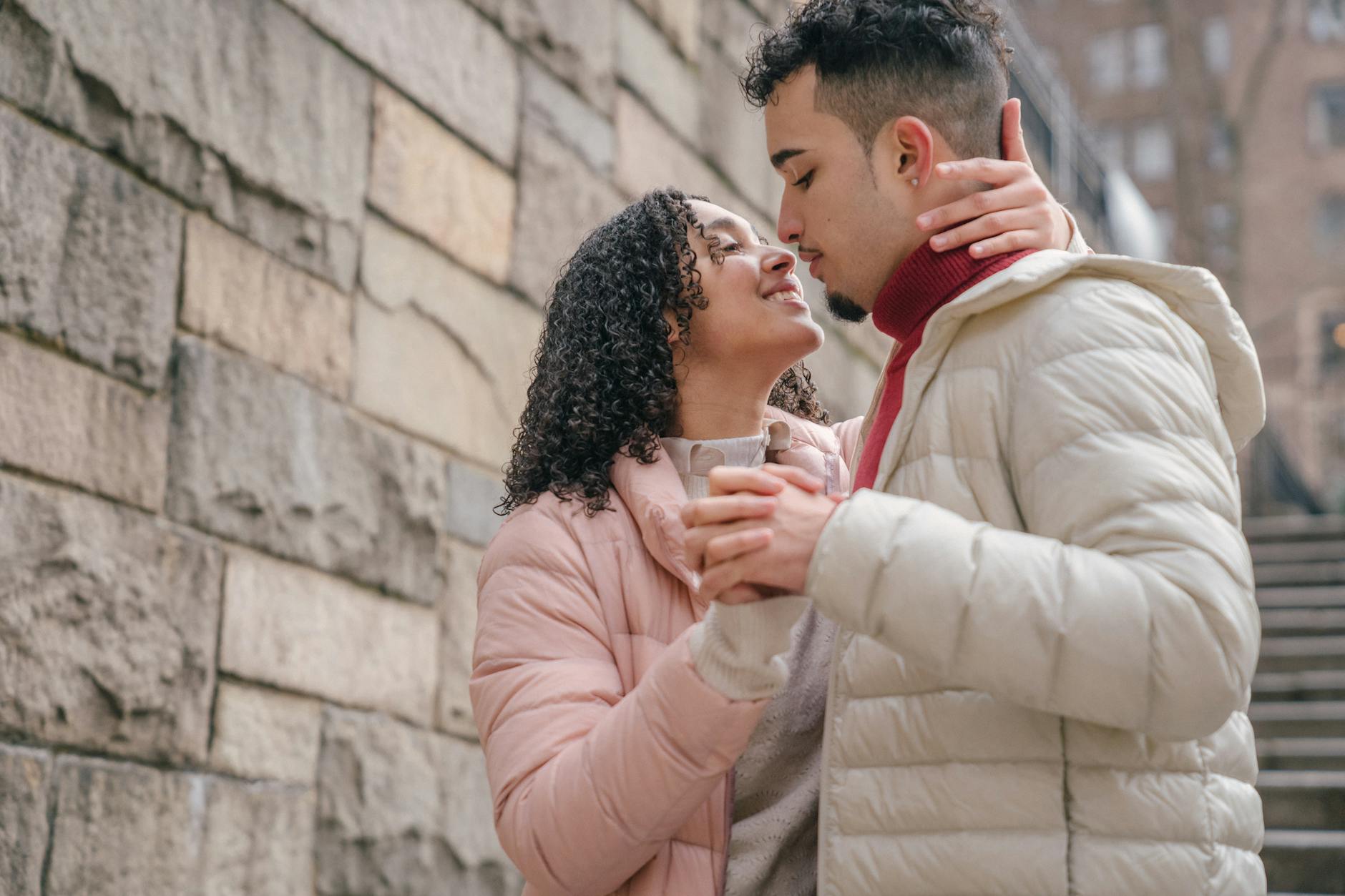 Side view of cheerful Hispanic couple holdings hand and looking at each other while hugging on street near stone wall - couple resolutions