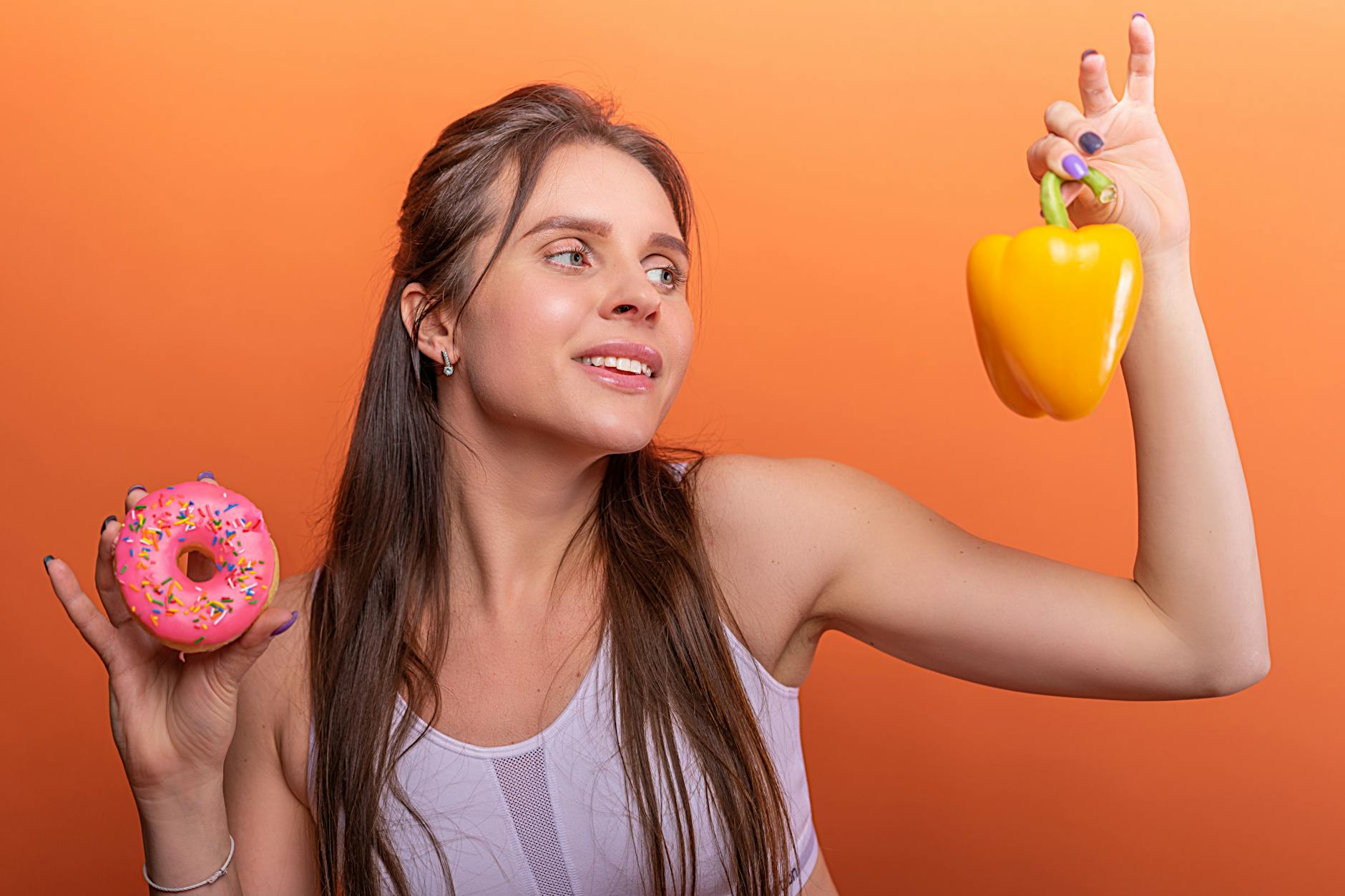 Woman in sports bra juggling choice between a donut and bell pepper on orange background. - diet for mental clarity