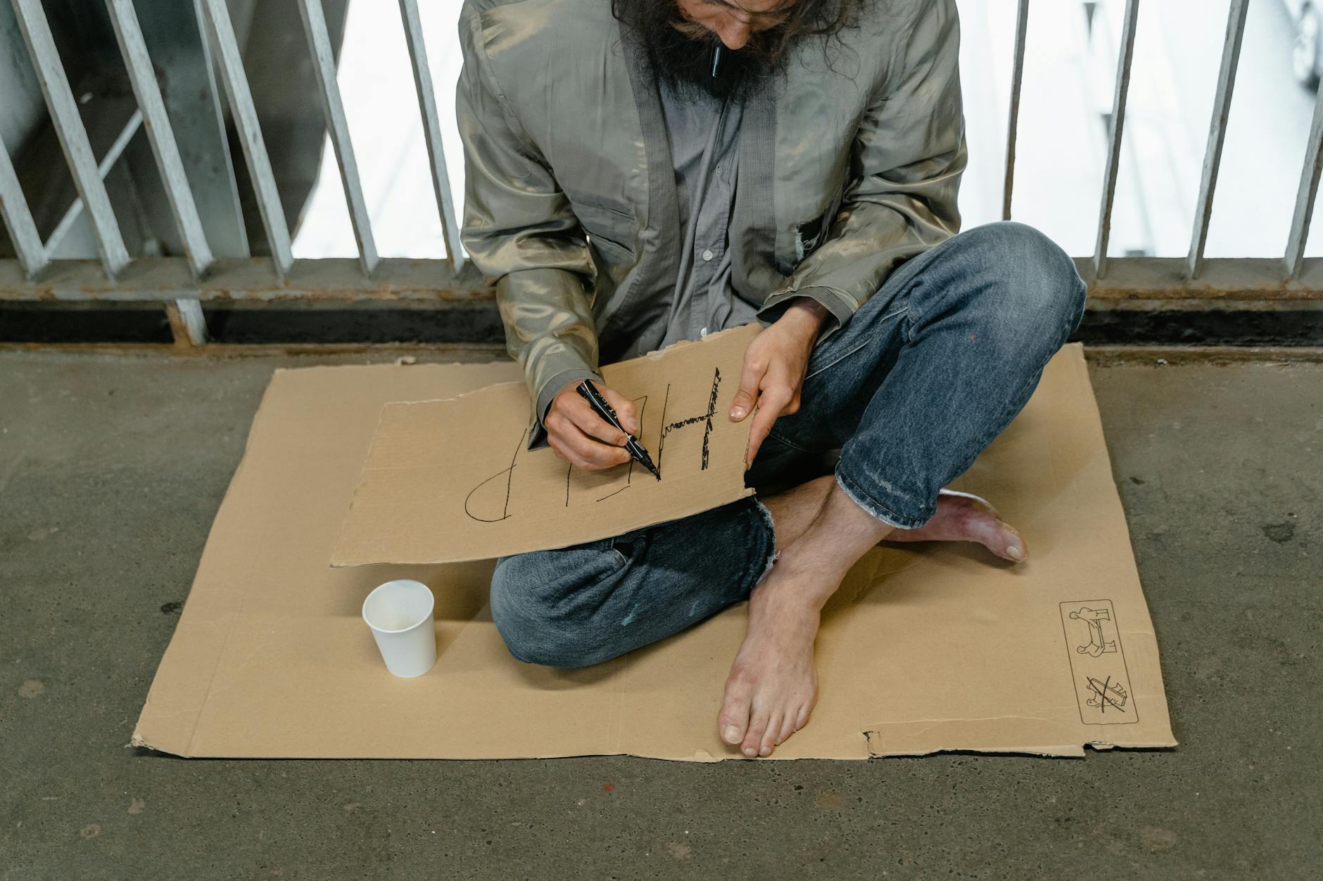 Person sitting on cardboard barefoot, writing a sign with a paper cup nearby. - express needs family