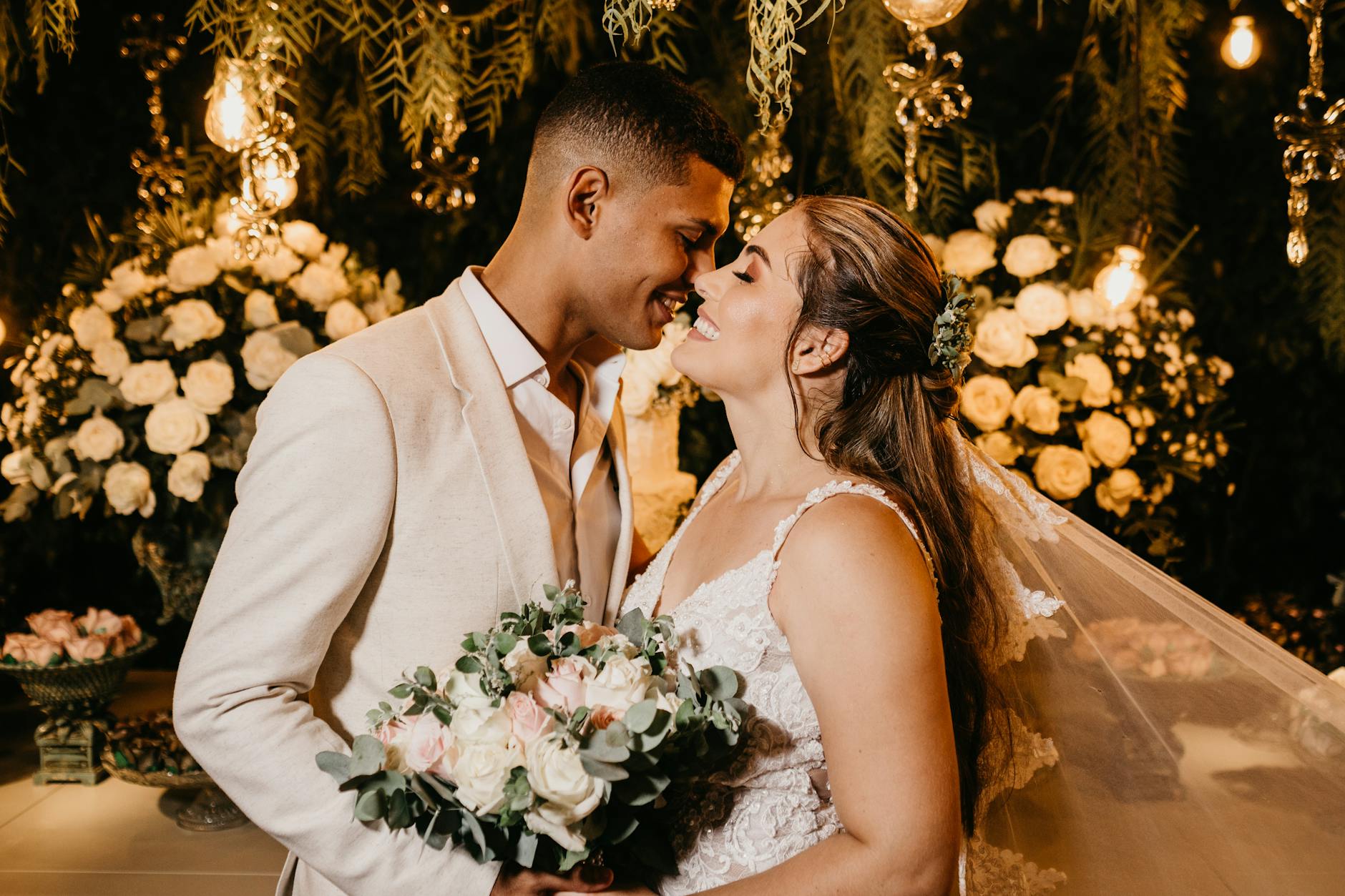 Cheerful young ethnic newlywed couple in elegant wedding apparel hugging with eyes closed against floral decorations in studio - expressing needs marriage