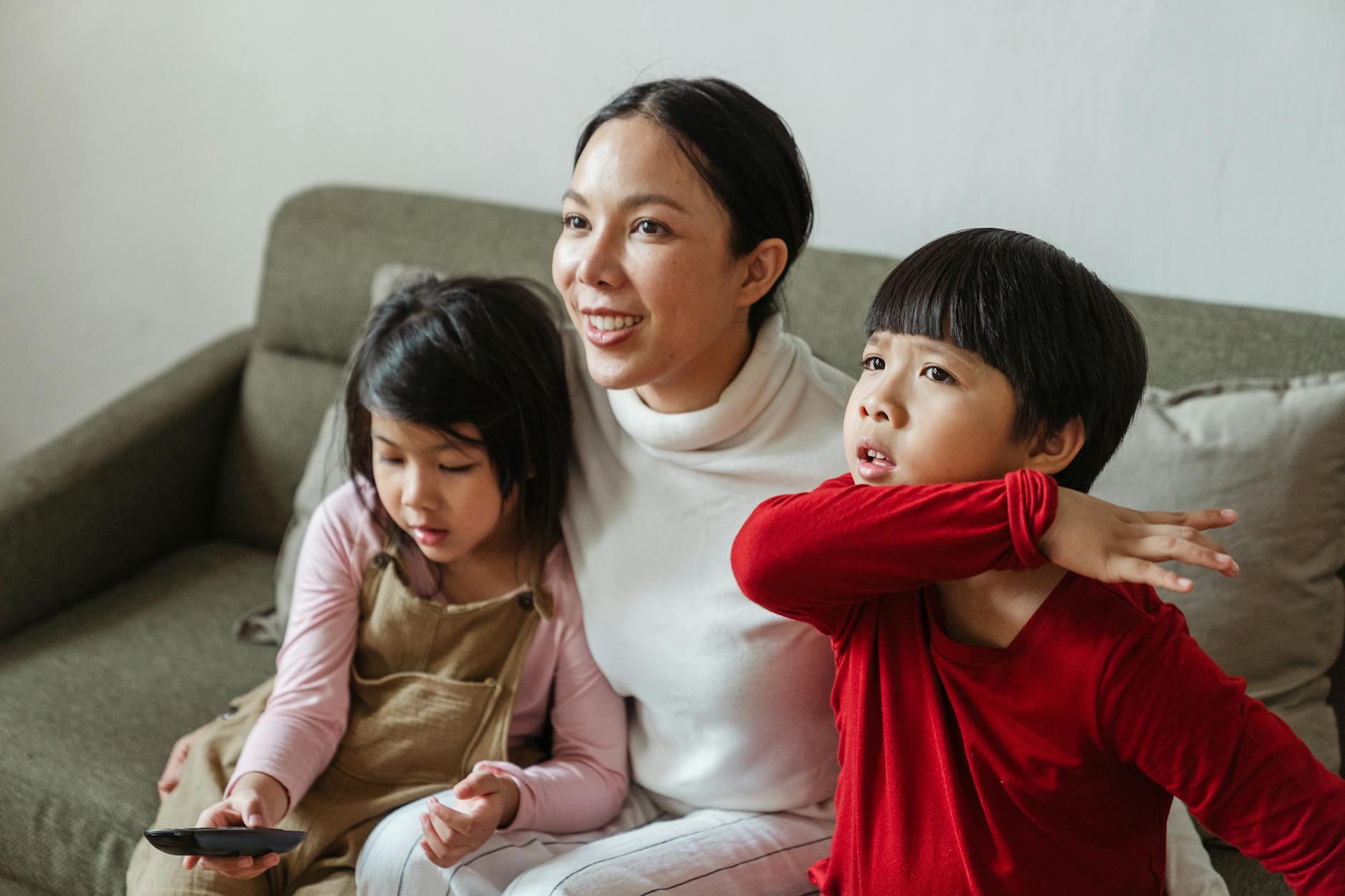 Positive Asian mom and little children watching interesting cartoon on TV while spending day together on comfortable couch - family guy sibling rivalry