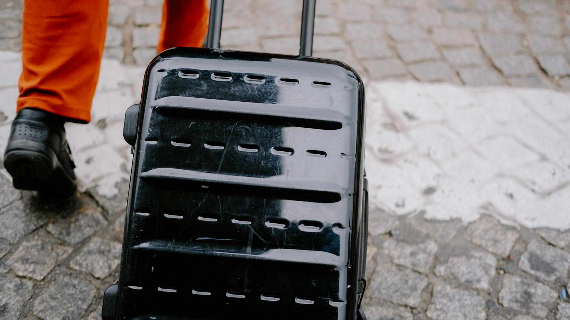 Close-up of a black suitcase being pulled on a cobblestone street by a person in orange pants. - letting go of baggage