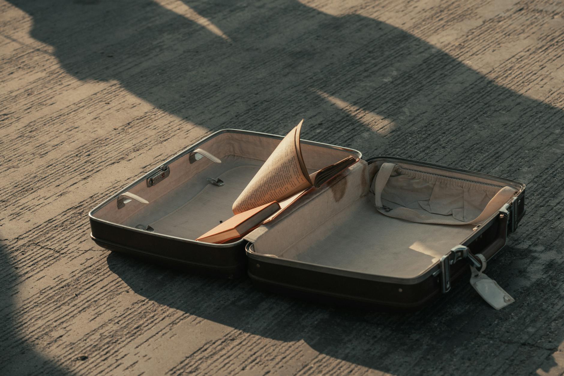 An open suitcase on a pavement with a book inside, casting shadows. - letting go of baggage