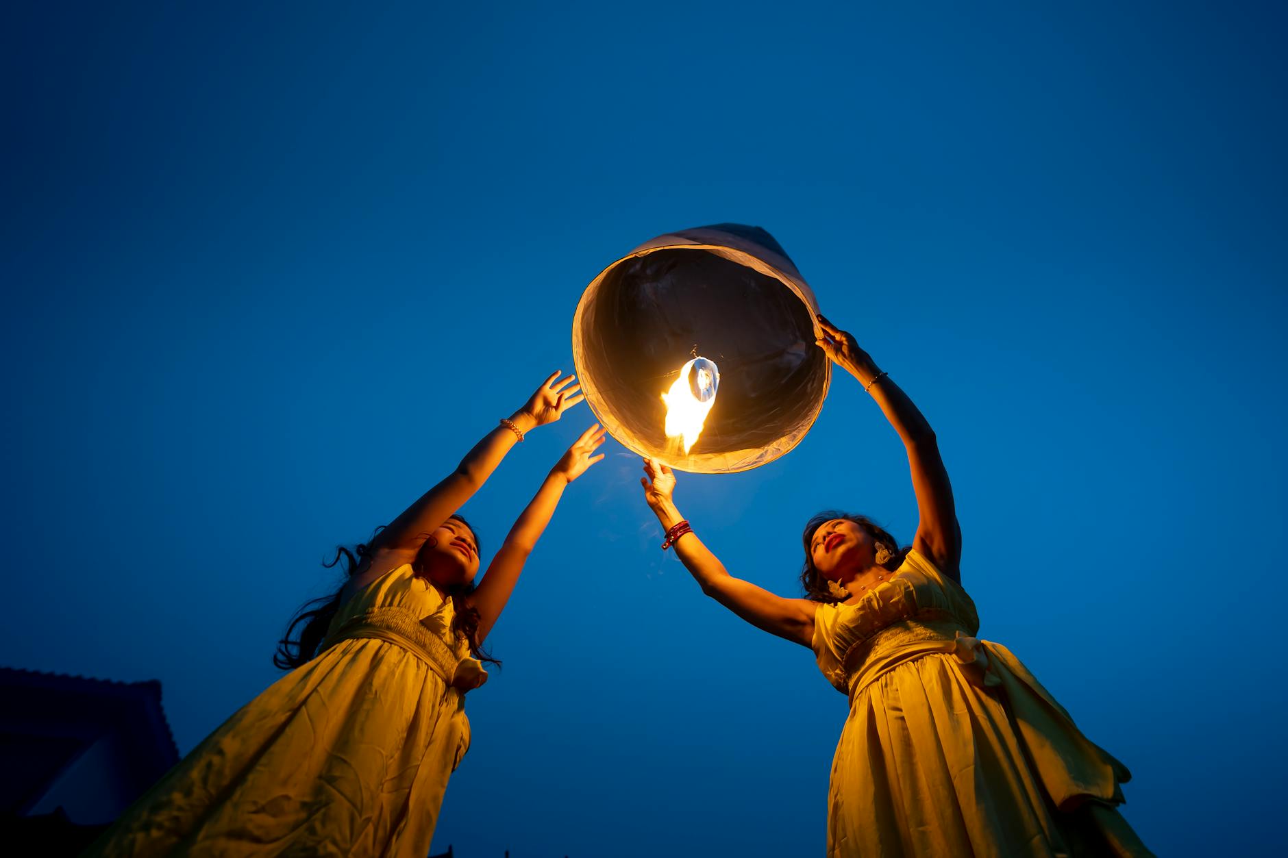Two women in yellow dresses releasing a glowing sky lantern into the evening sky, symbolizing hope and celebration. - letting go resentment