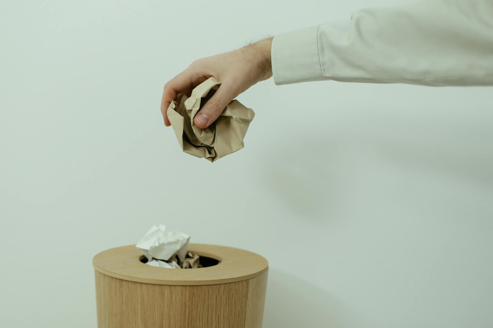 A person's hand disposes of crumpled paper into a wooden trash bin indoors. - mental health decluttering