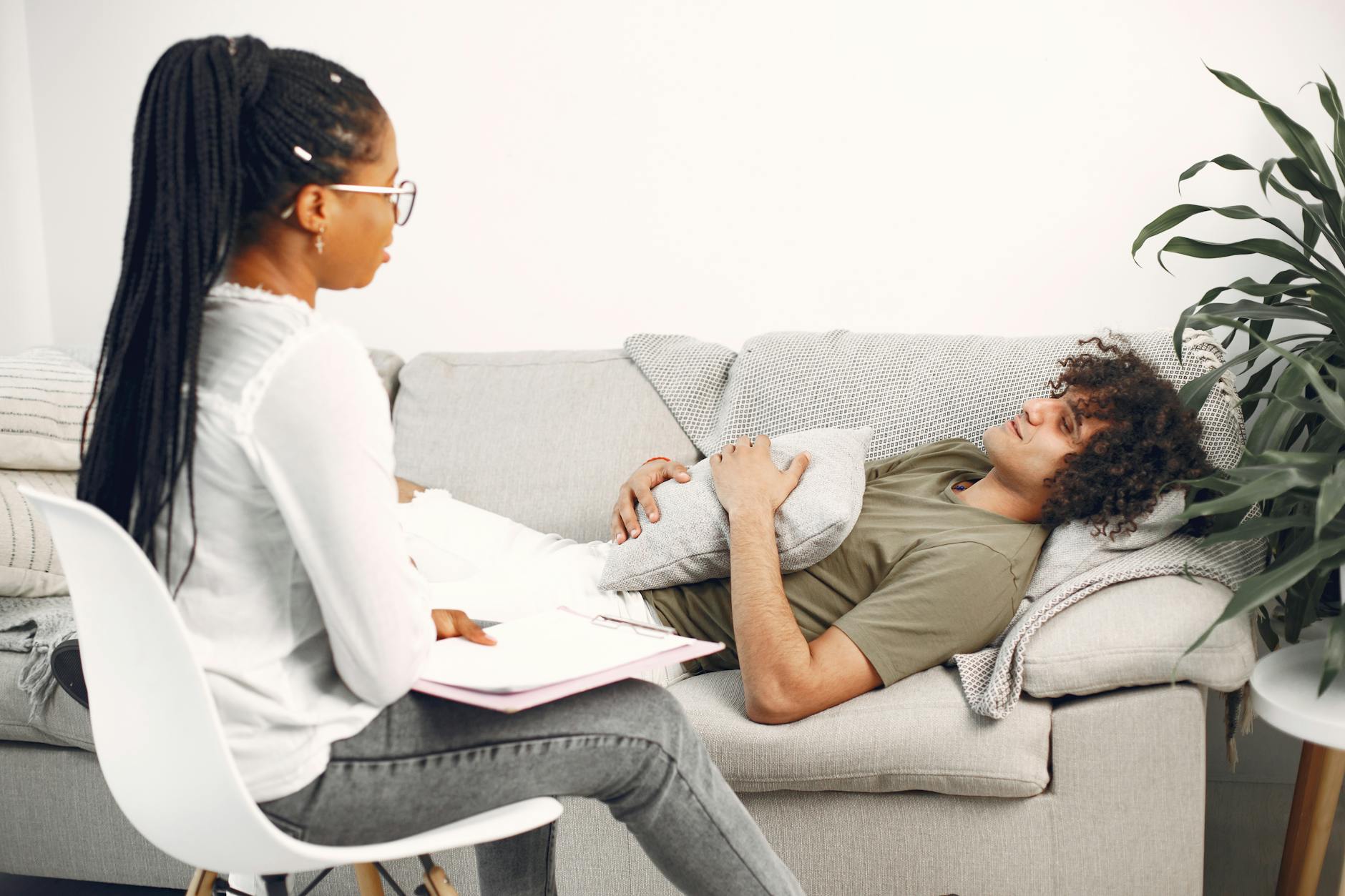 A man in therapy session, lying on a couch, speaking with a female therapist taking notes. - mental well-being therapy