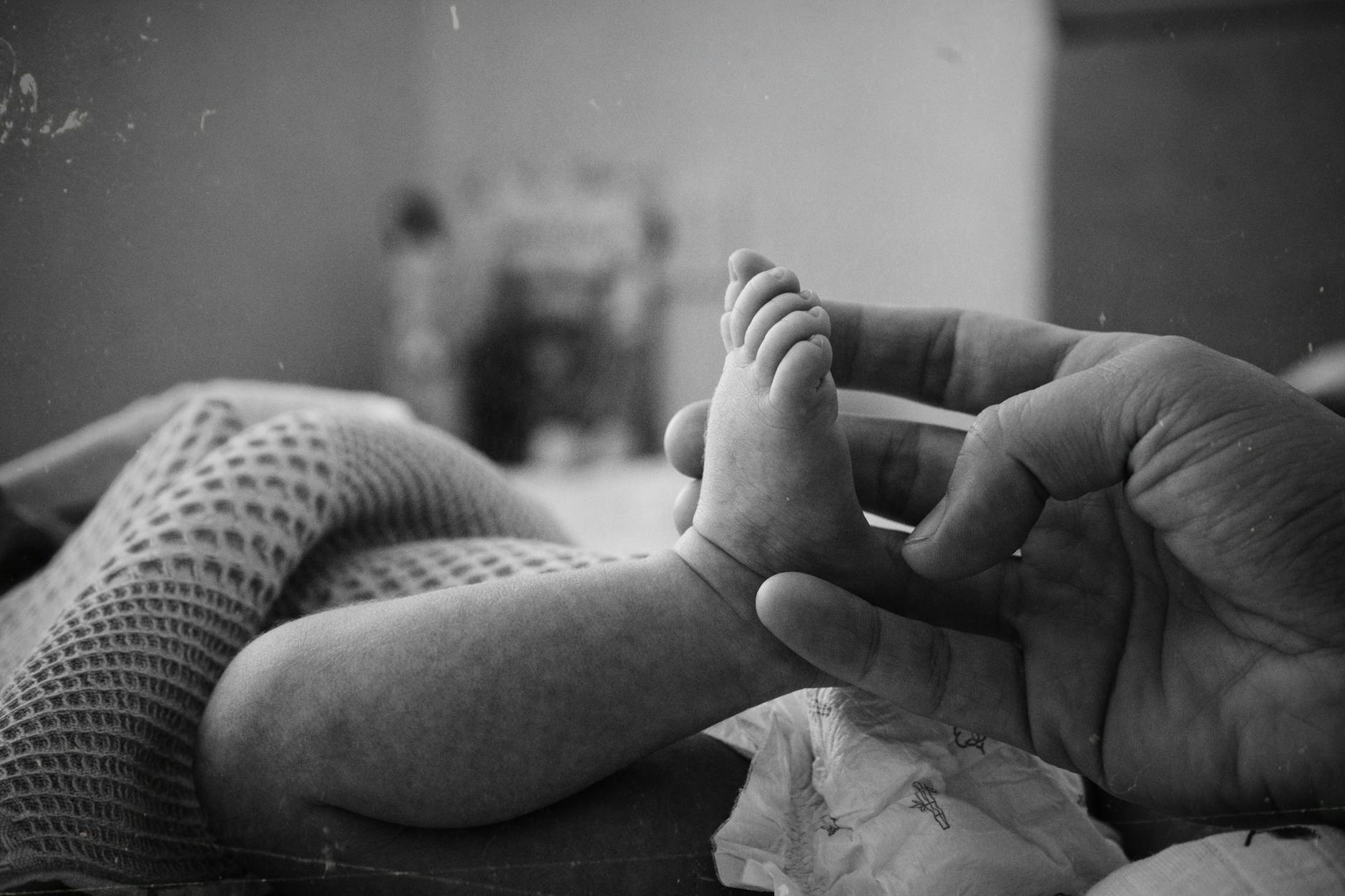 Tender black and white image of a newborn's foot being gently held by an adult. - new parent exhaustion