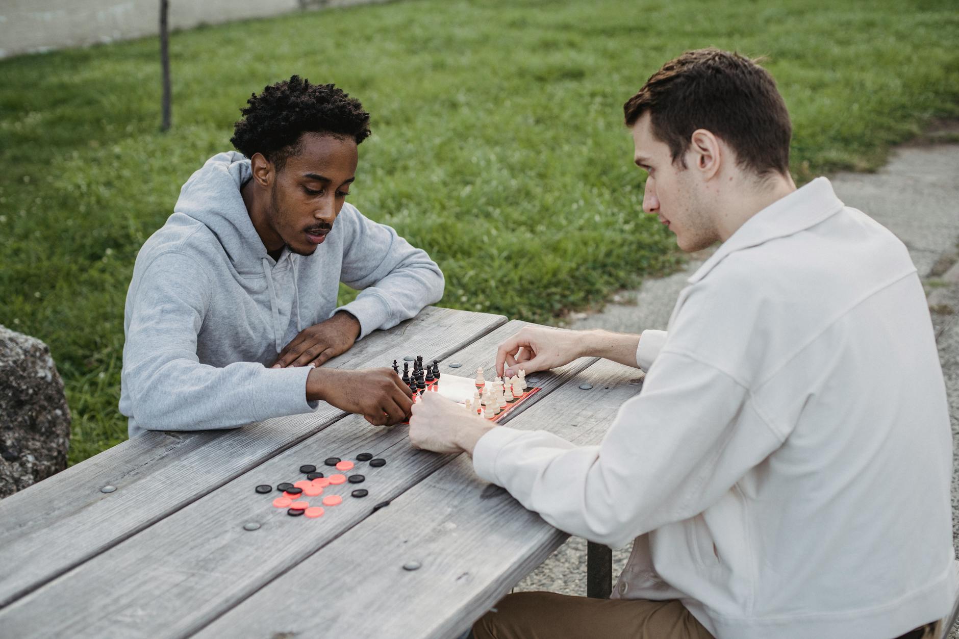 Concentrated young multiethnic male friends in casual clothes sitting at table in park and playing chess game - outdoor social skills