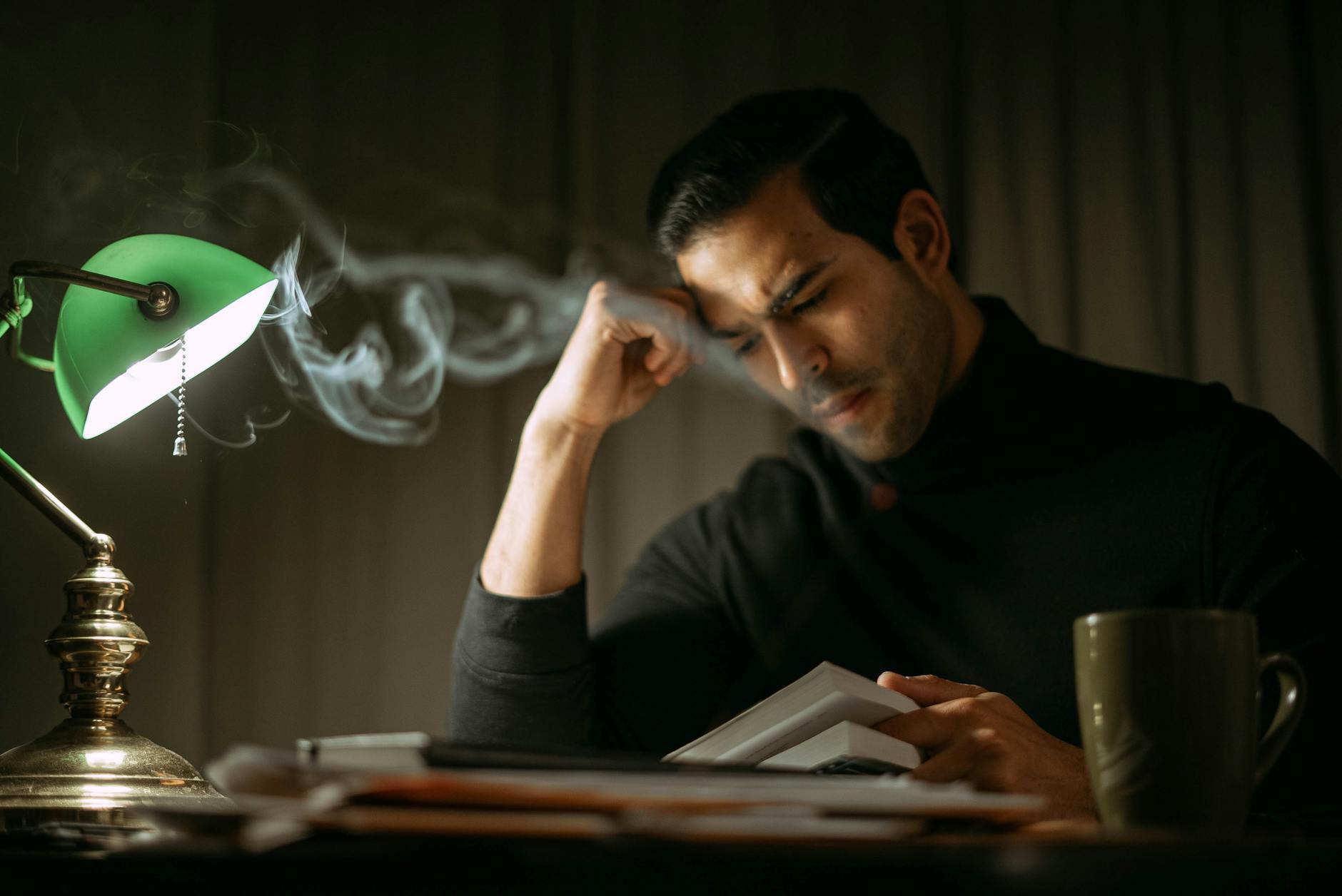 Frowning handsome elegant male leaning on hands while reading book at table with retro lamp in fume - overcome mental fog