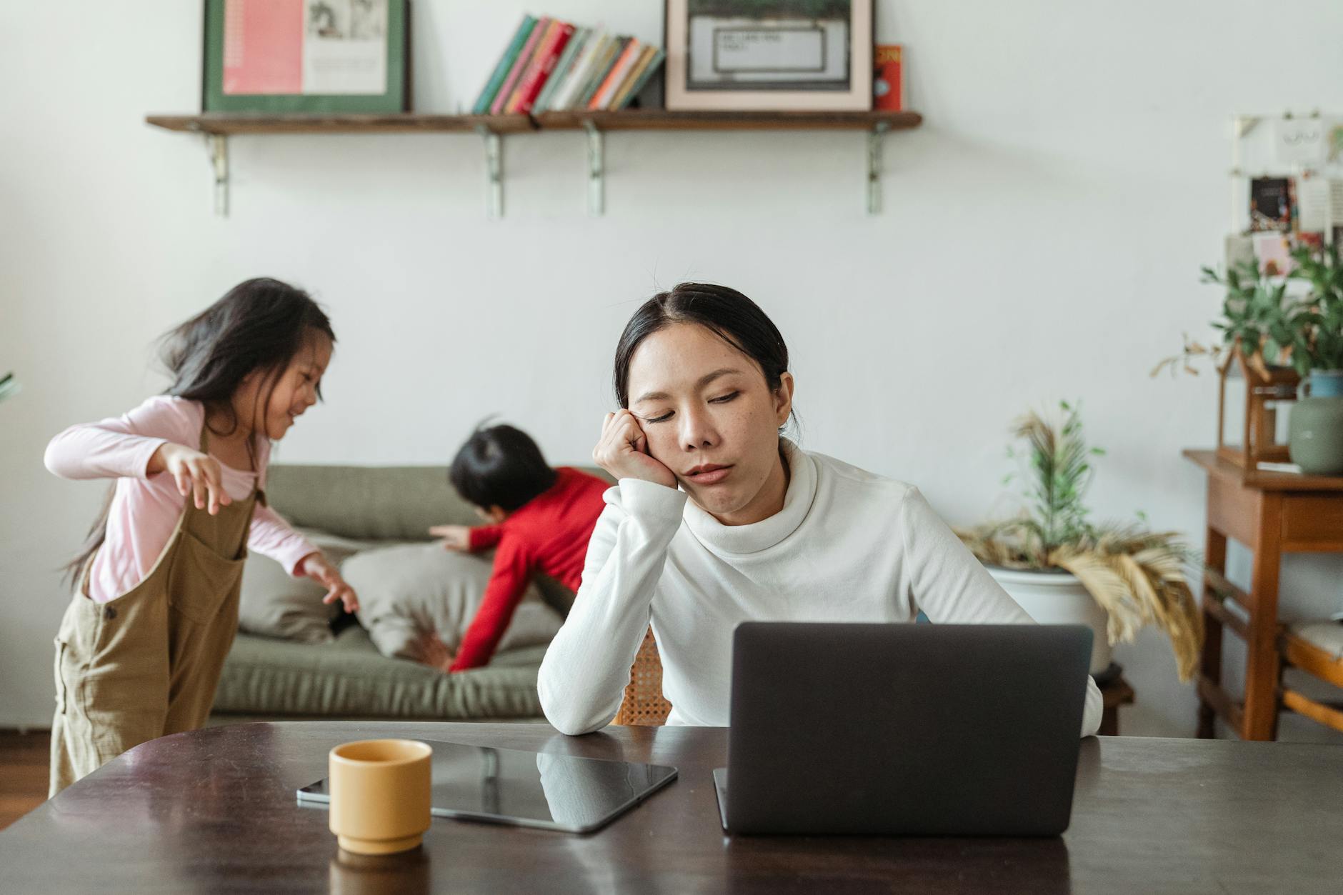 Overworked young Asian female freelancer sitting at table with laptop and tablet against active disturbing children - parent stress relief