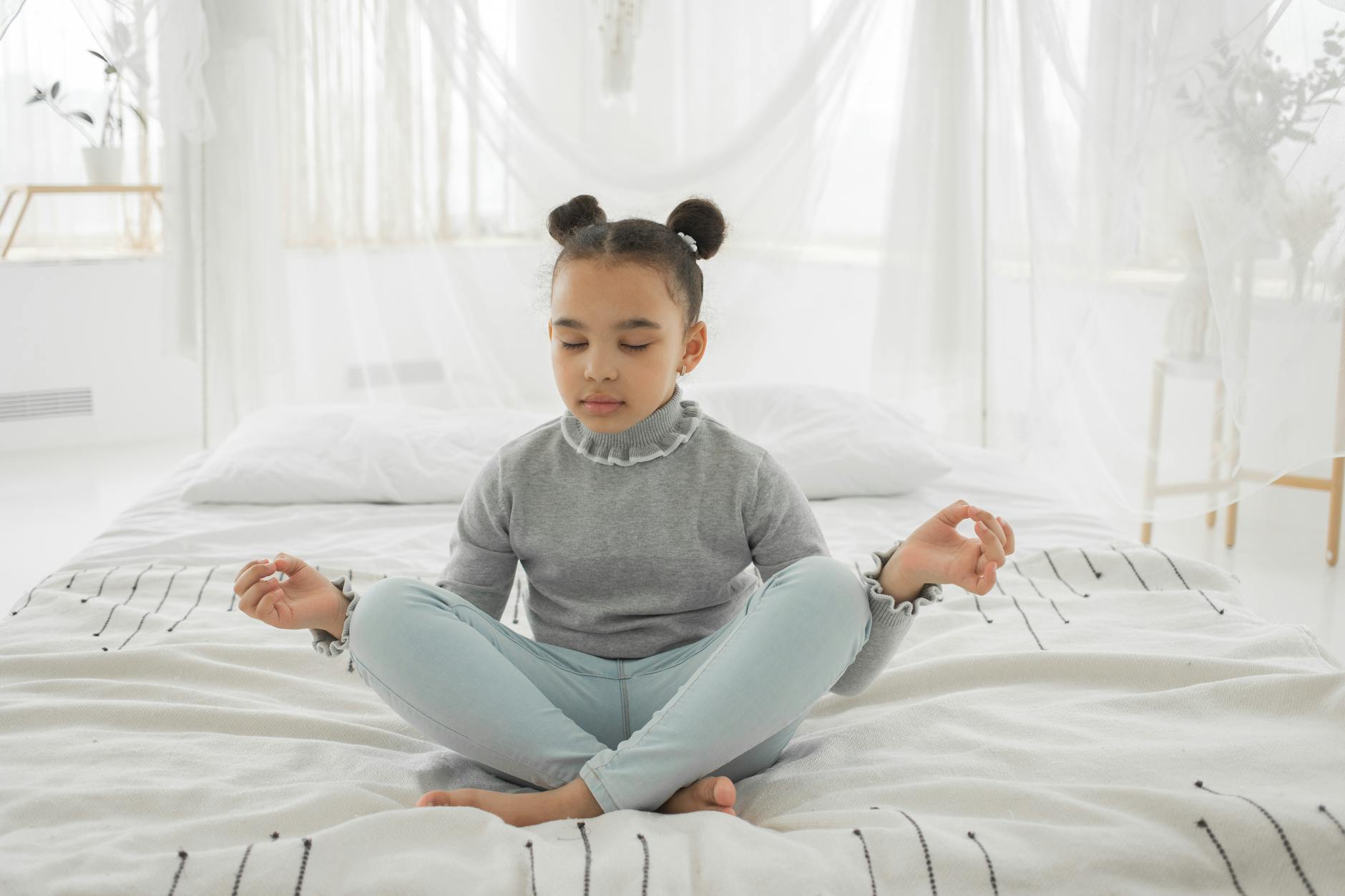 Full body of adorable little ethnic girl with pig buns in casual clothes sitting on soft bed with crossed legs while meditating with closed eyes in light bedroom - parent stress relief