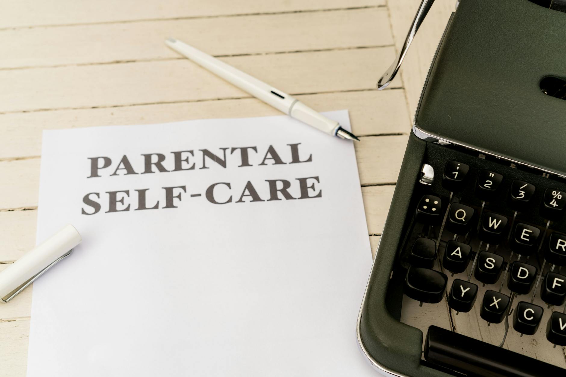 Close-up of paper with 'Parental Self-Care' beside a typewriter, pen, and clip on a wooden desk. - parent stress relief