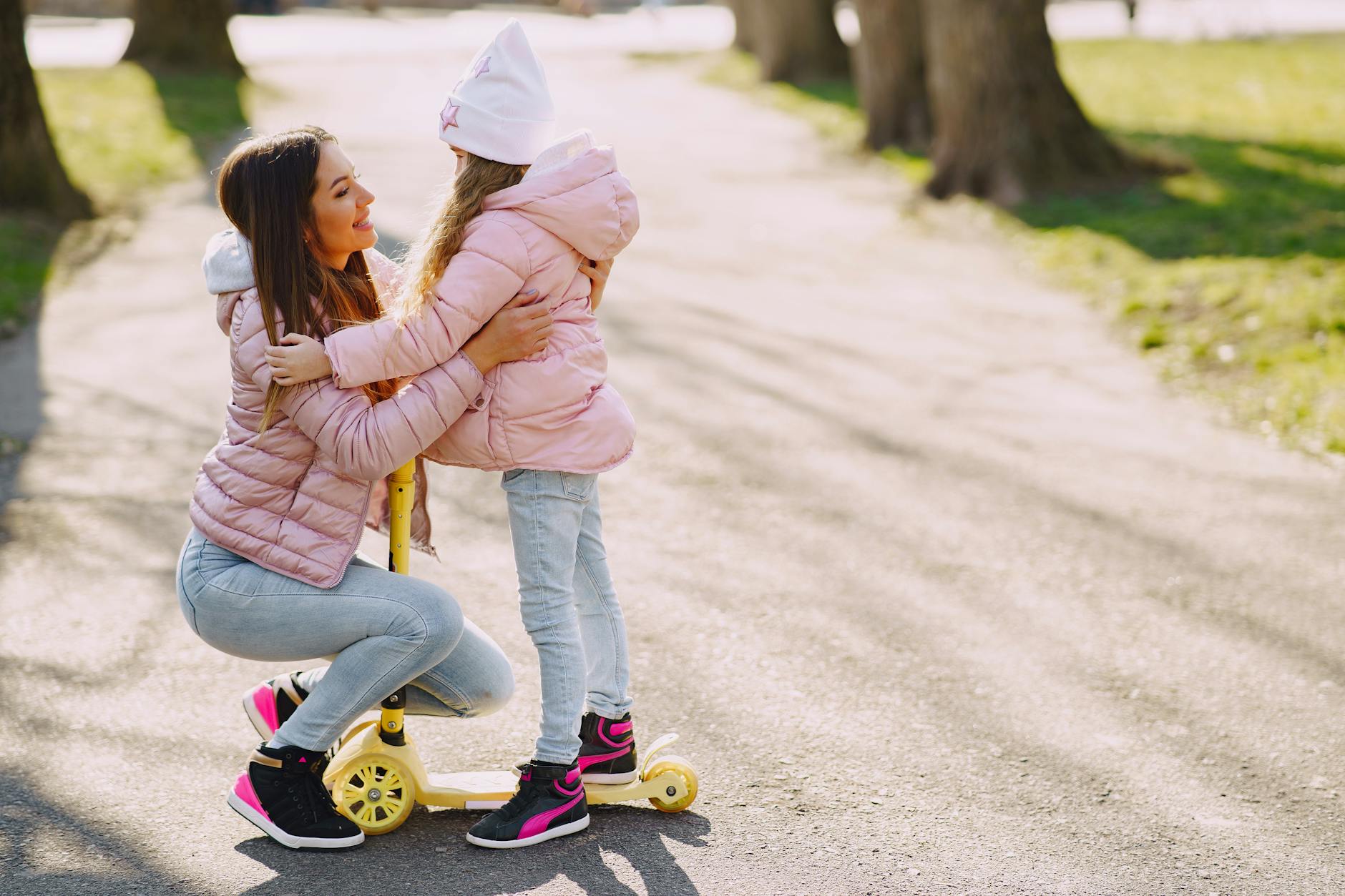 Side view of little girl in casual warm wear hugging mother while riding kick scooter on asphalt alley in city park and looking to each other - parental burnout spring