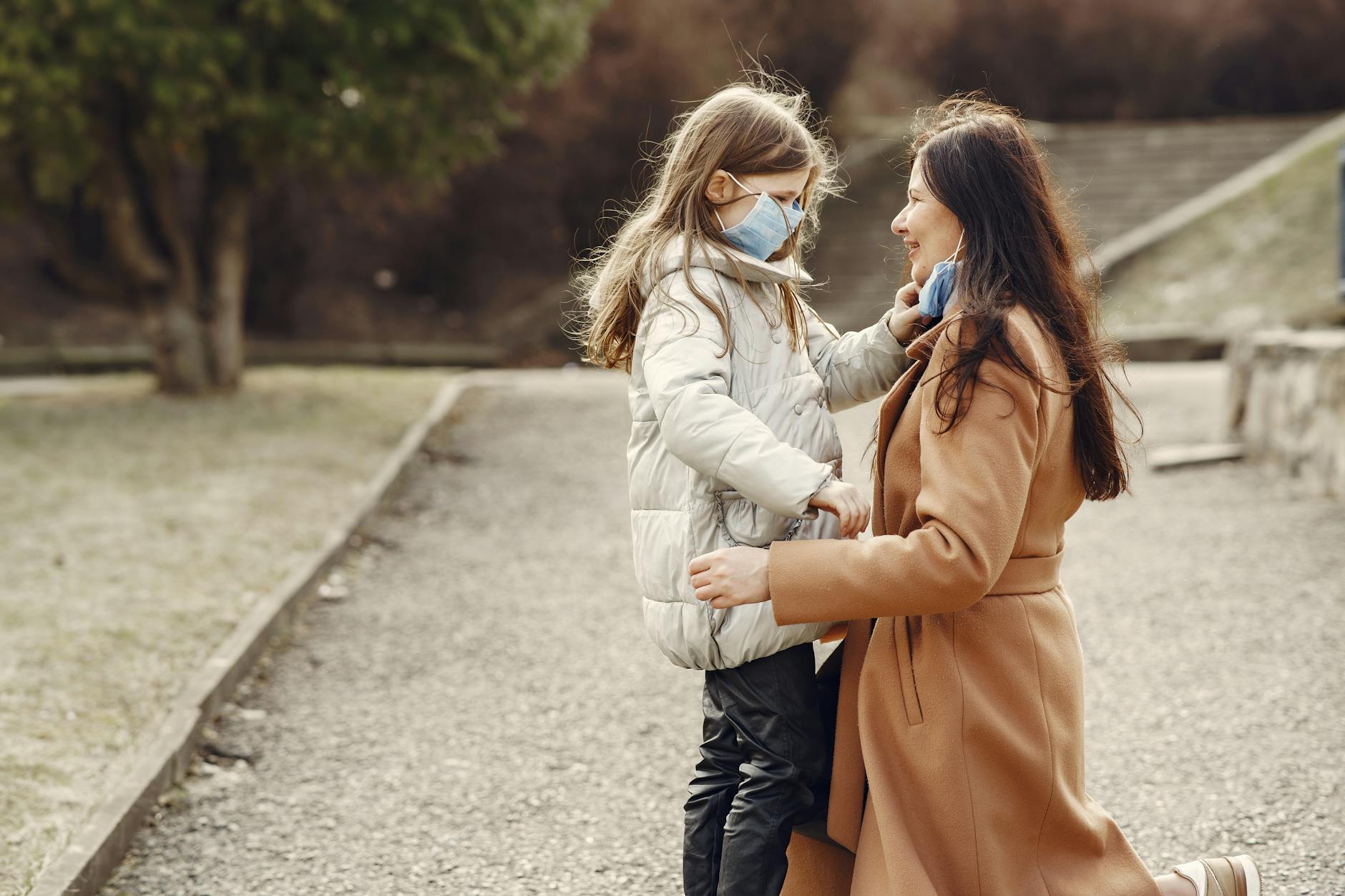 Side view of little girl in warm clothes and mask taking off mask from face of smiling woman in warm coat during stroll in park - post pandemic family