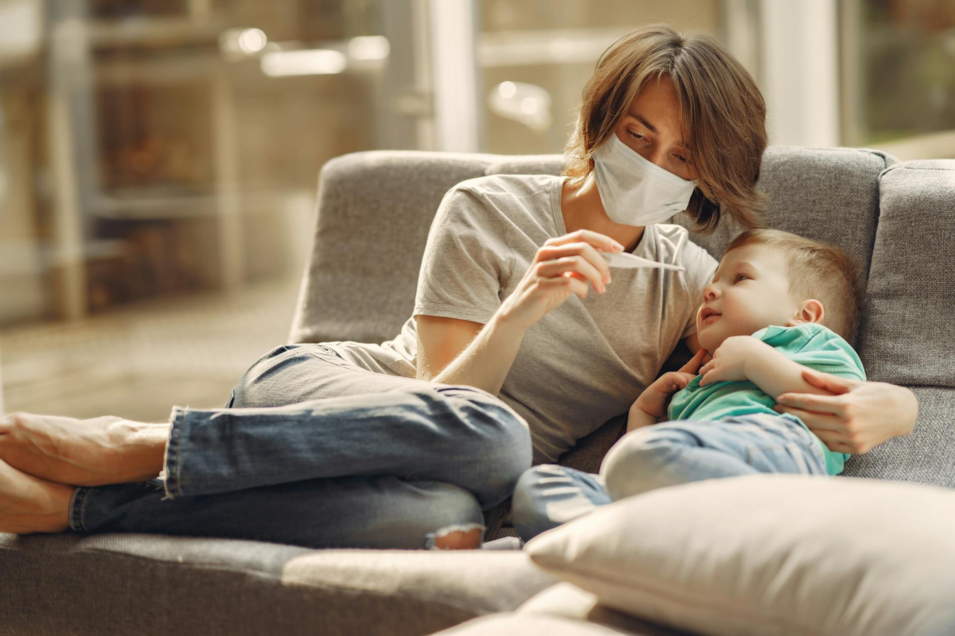 A caring mother checks her son's temperature while they relax on a sofa, showcasing parent-child affection and health care. - post pandemic family