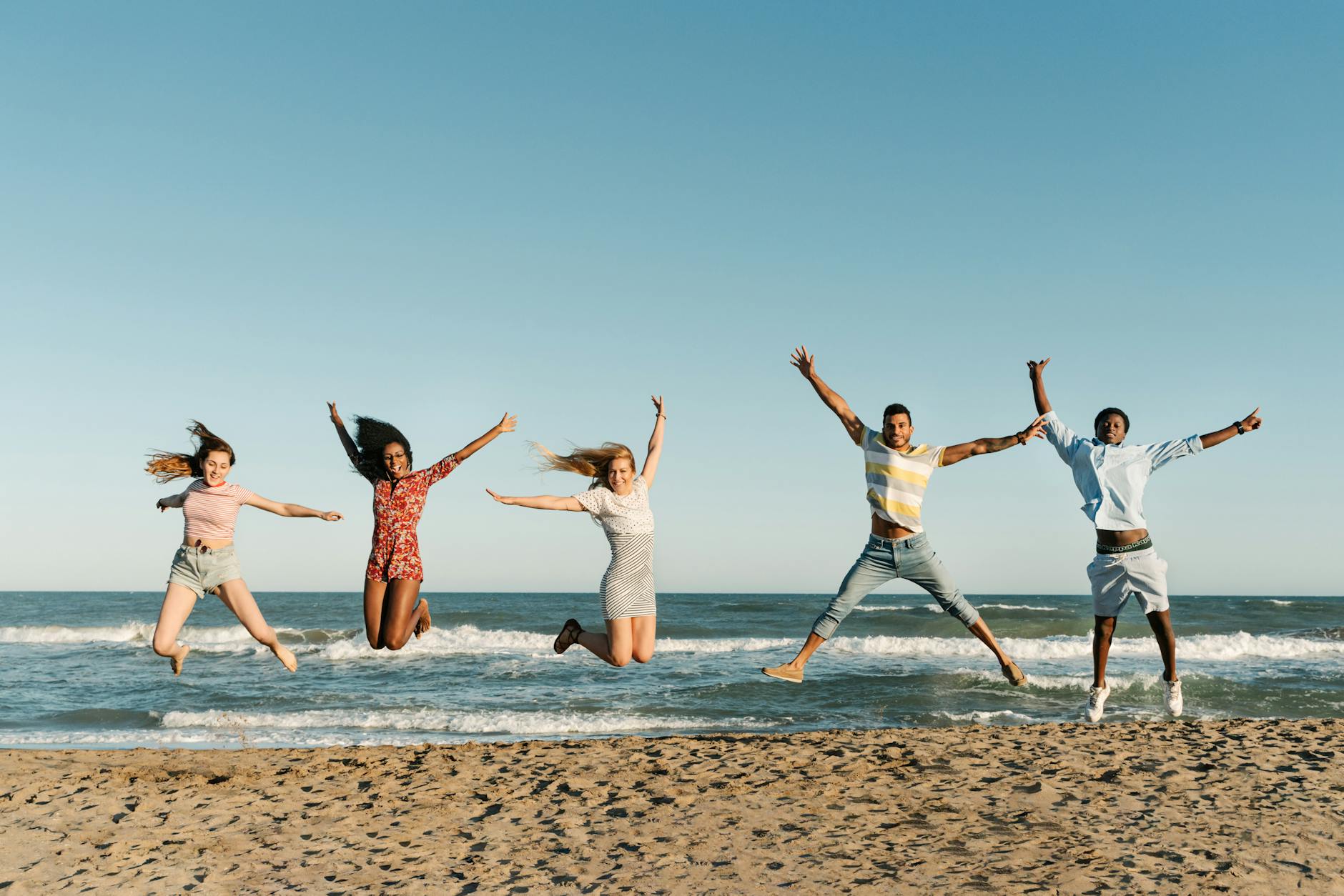 Happy diverse tourists jumping with raised arms over sandy ocean shore under blue sky during summer trip while looking at camera - post spring break reconnect