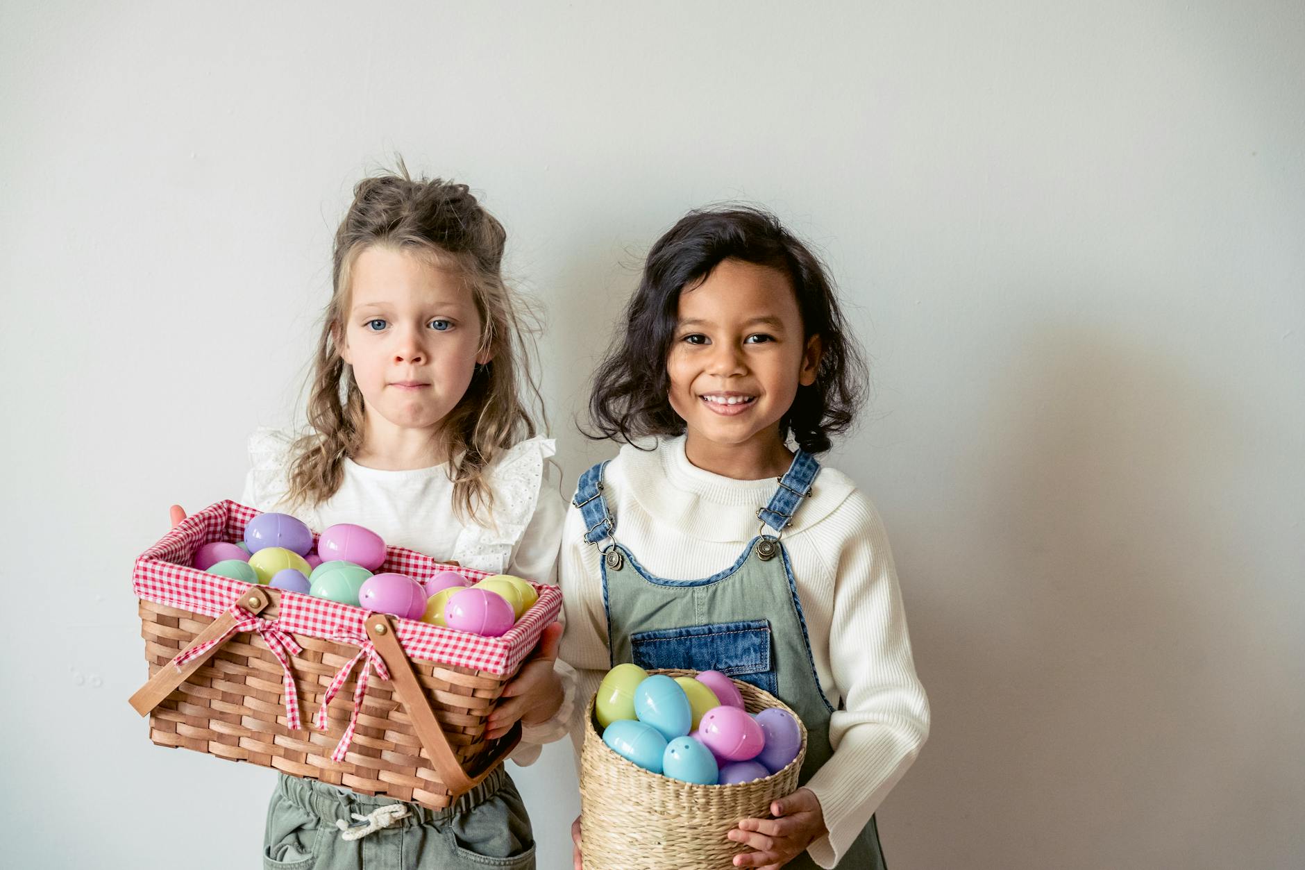 Delighted diverse girls with wicker baskets full of colored eggs standing on white background during Easter holiday celebration in light room - preschool spring changes