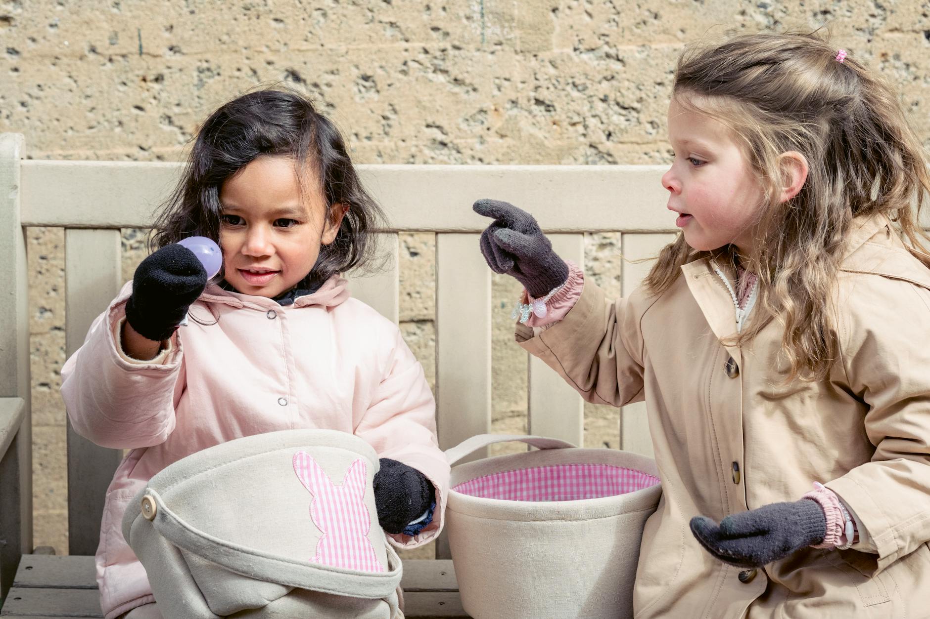 Adorable multiracial girls in outerwear sitting on bench with baskets while playing egg hunt game on street during Easter holiday - preschool spring changes