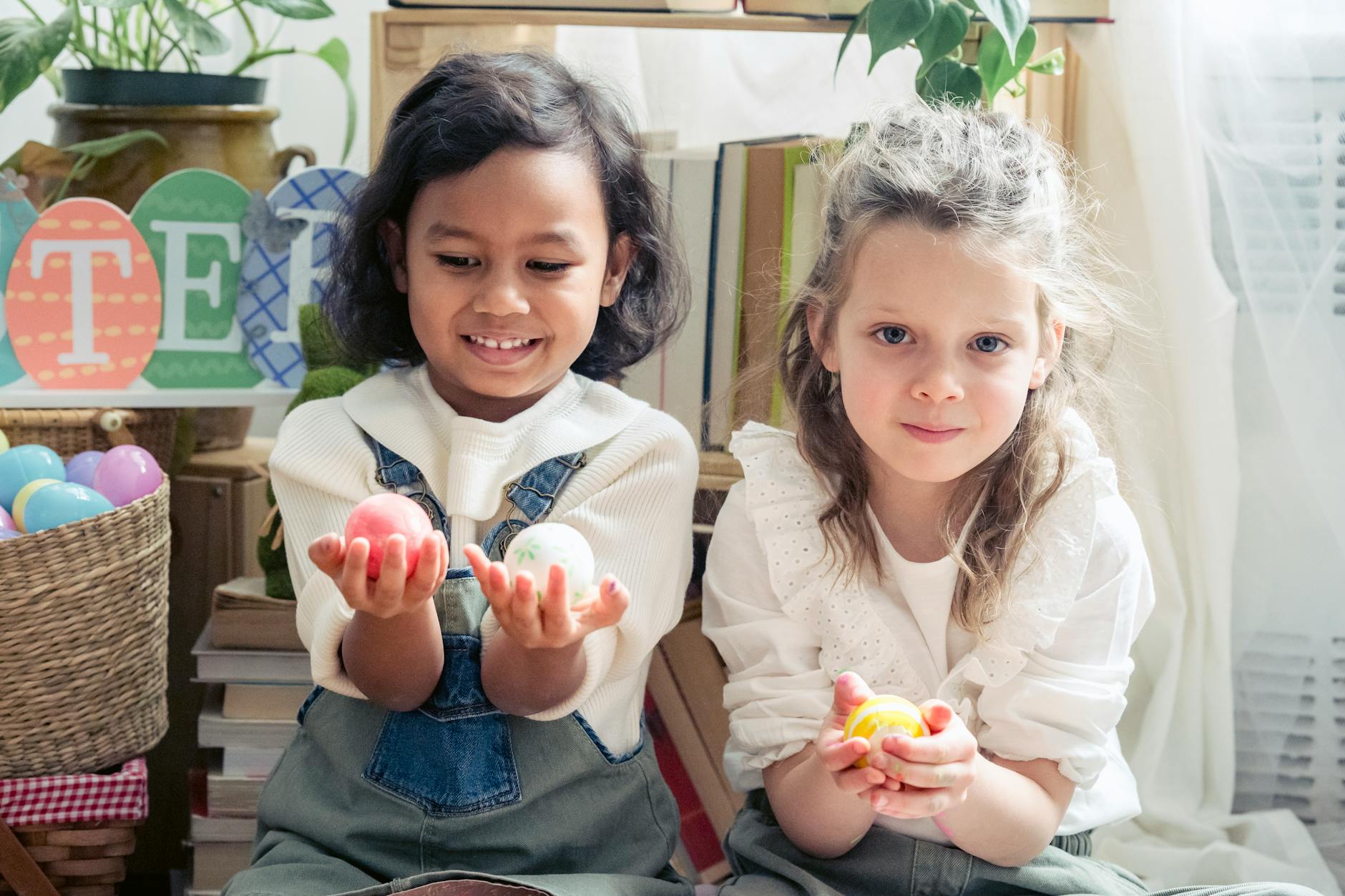 Cute little diverse girls with colorful eggs sitting in light room with creative decorations during Easter holiday celebration at home - preschool spring schedule