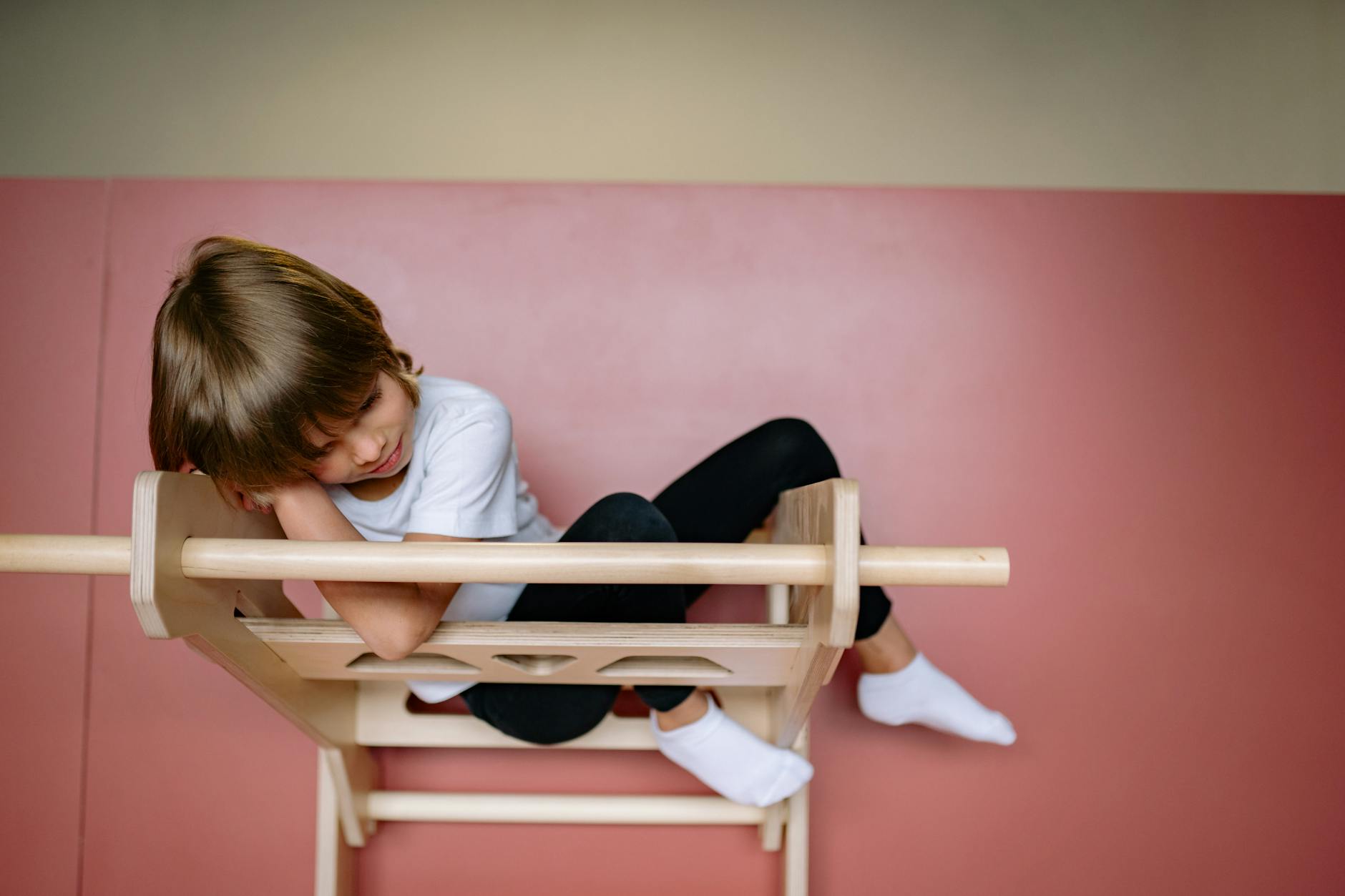A young child with brown hair in a t-shirt and leggings reclines on wooden gymnastic equipment indoors. - preschooler spring adjustment