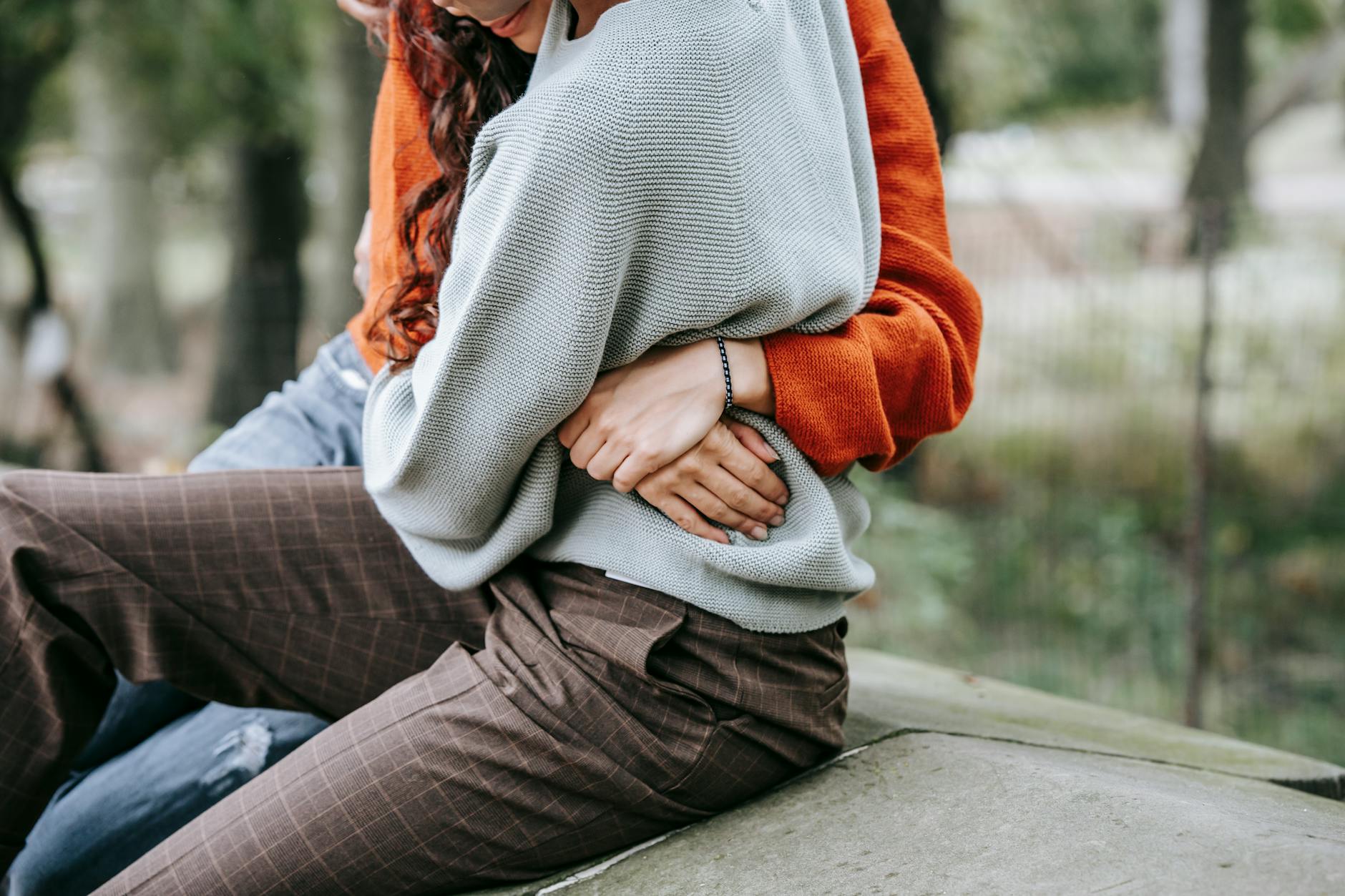 Crop anonymous women hugging each other while sitting on stone surface together in casual clothes in daytime - reconnect with partner