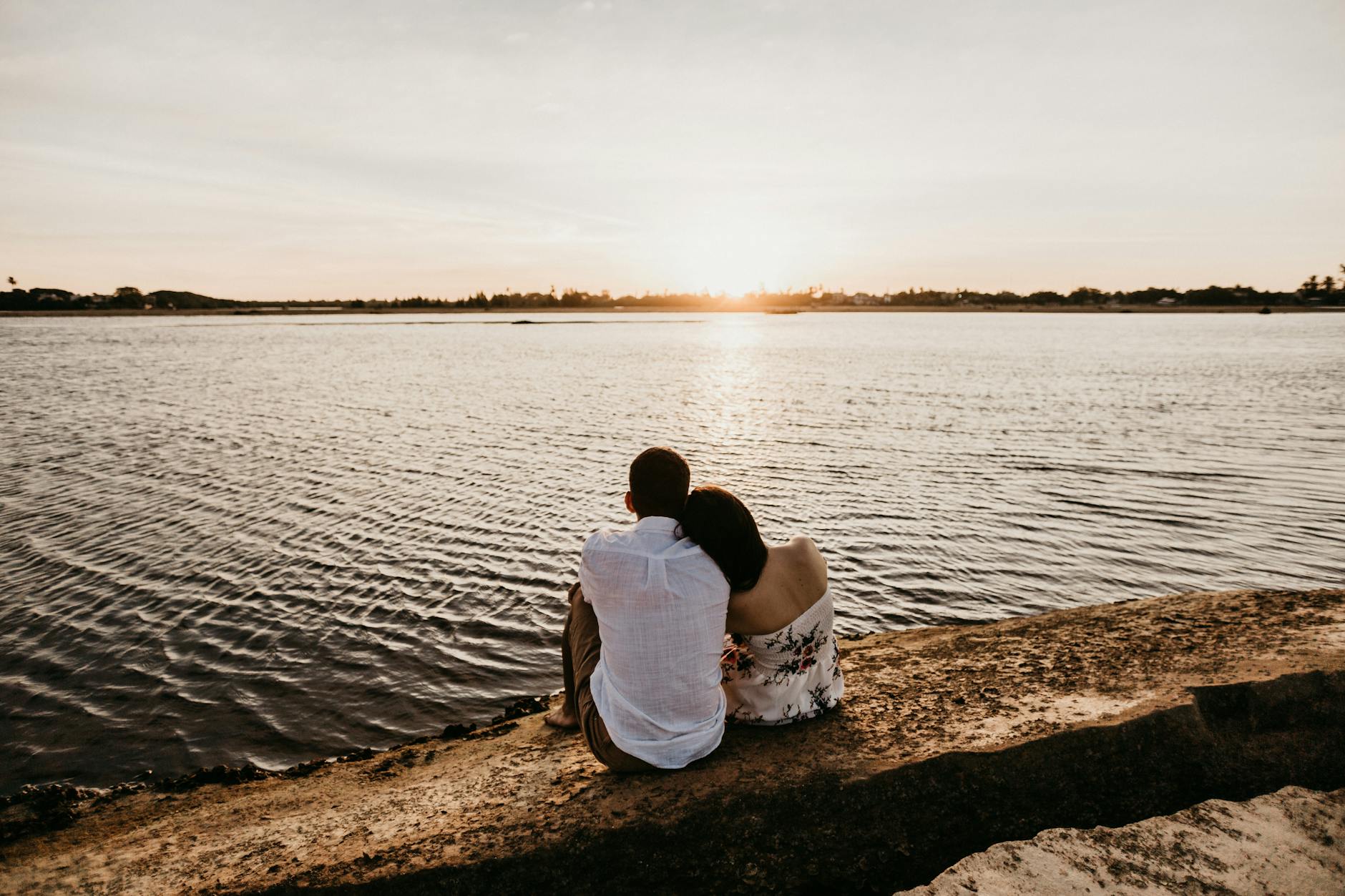 Back view unrecognizable female resting head on boyfriends shoulder while sitting together on stony river coast during romantic sunset - reconnect with partner