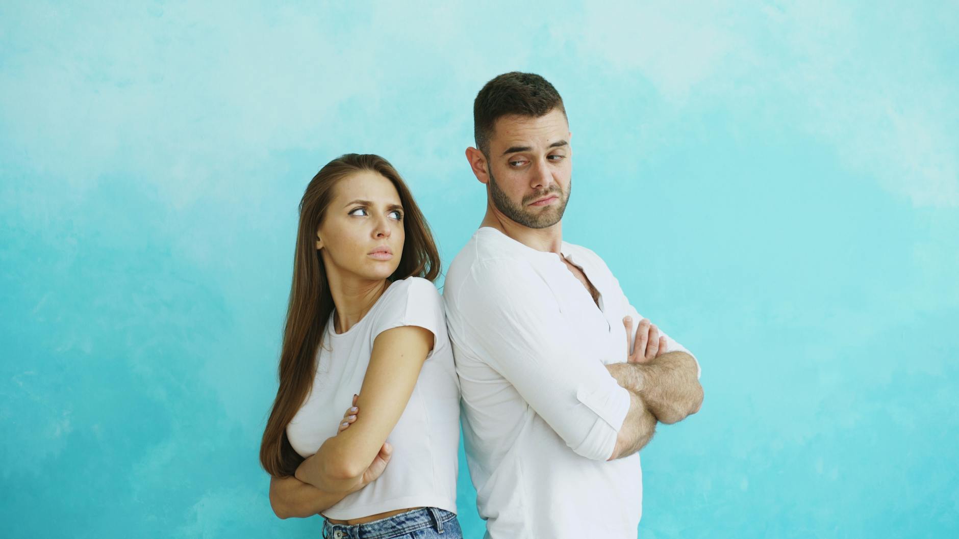 Young couple stands back to back with crossed arms against blue background. - relationship resentment