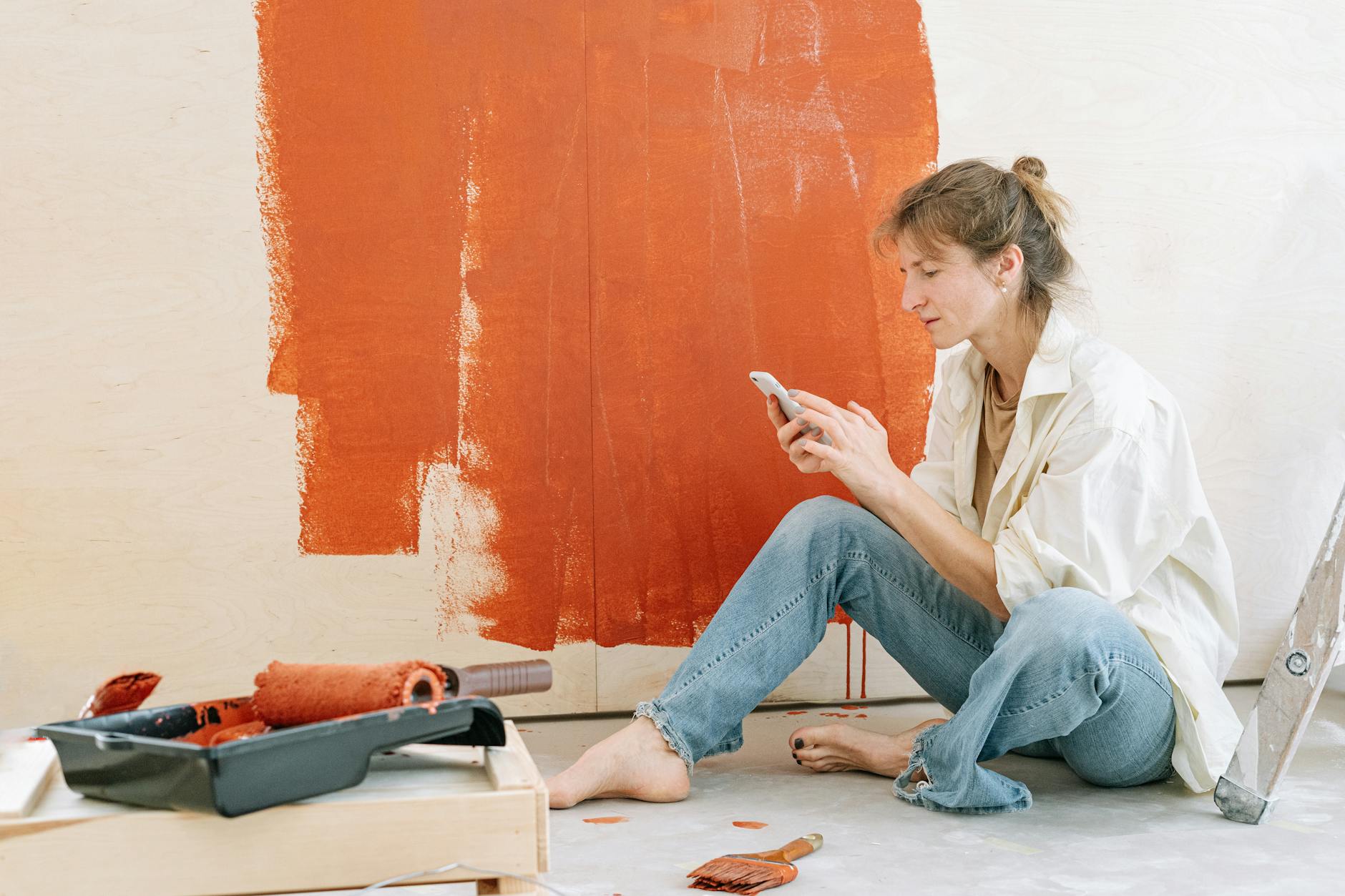 A woman takes a break from renovating by checking her phone while painting a wall. - renovation communication needs