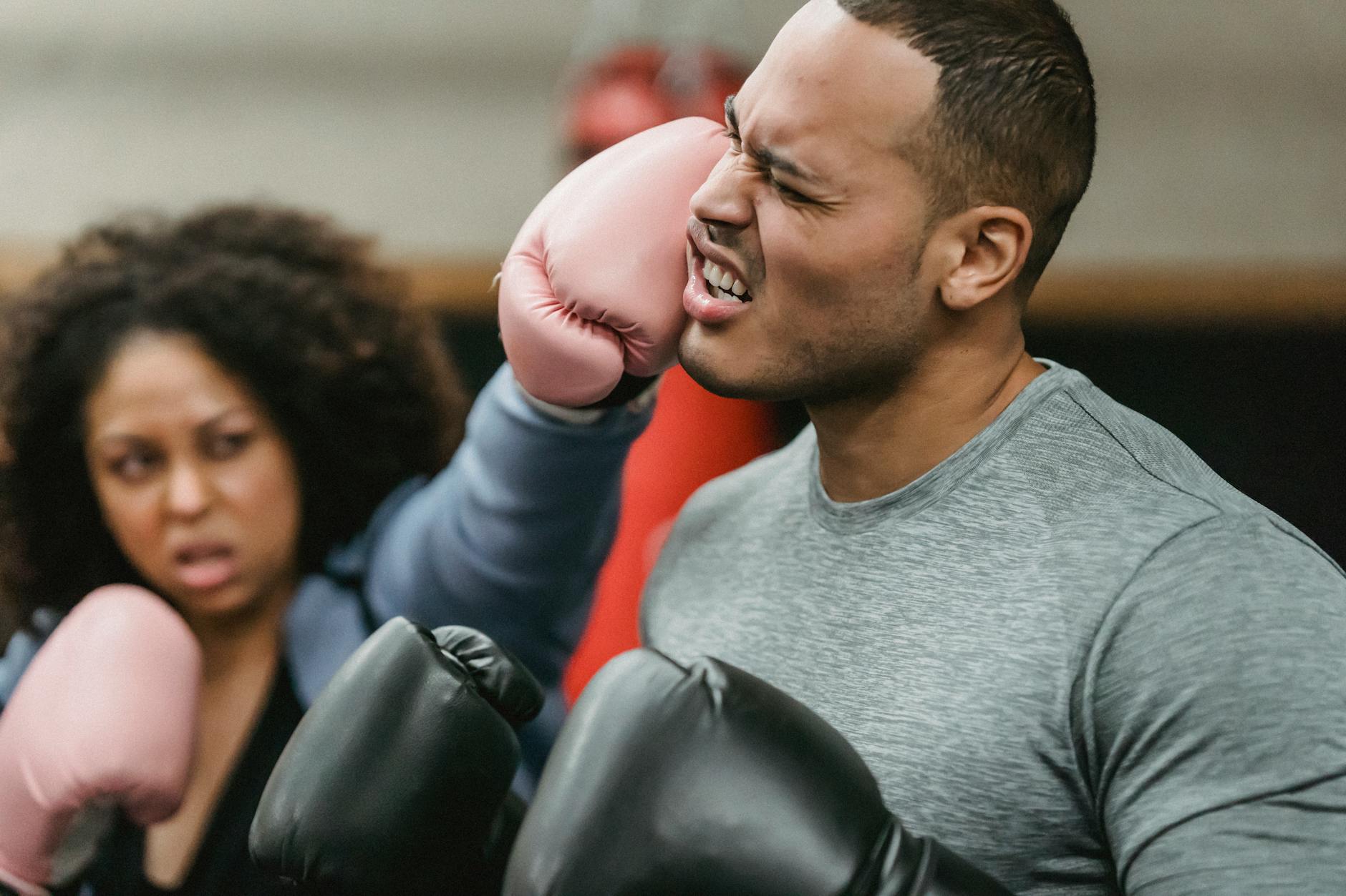 Side view of young muscular ethnic male trainer in sportswear and boxing gloves receiving heavy punch on face from serious young African American female during workout - repairing trust after fight
