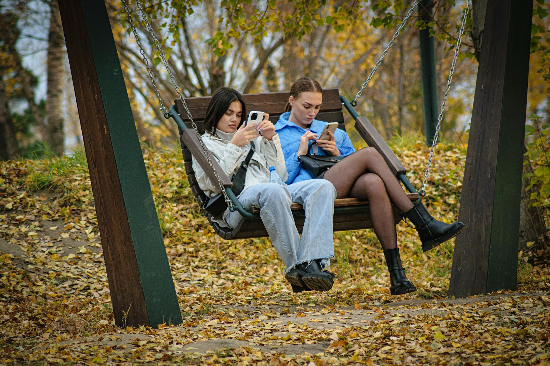 Two young women sitting on a swing using smartphones, surrounded by autumn leaves in a park. - seasonal mood swings