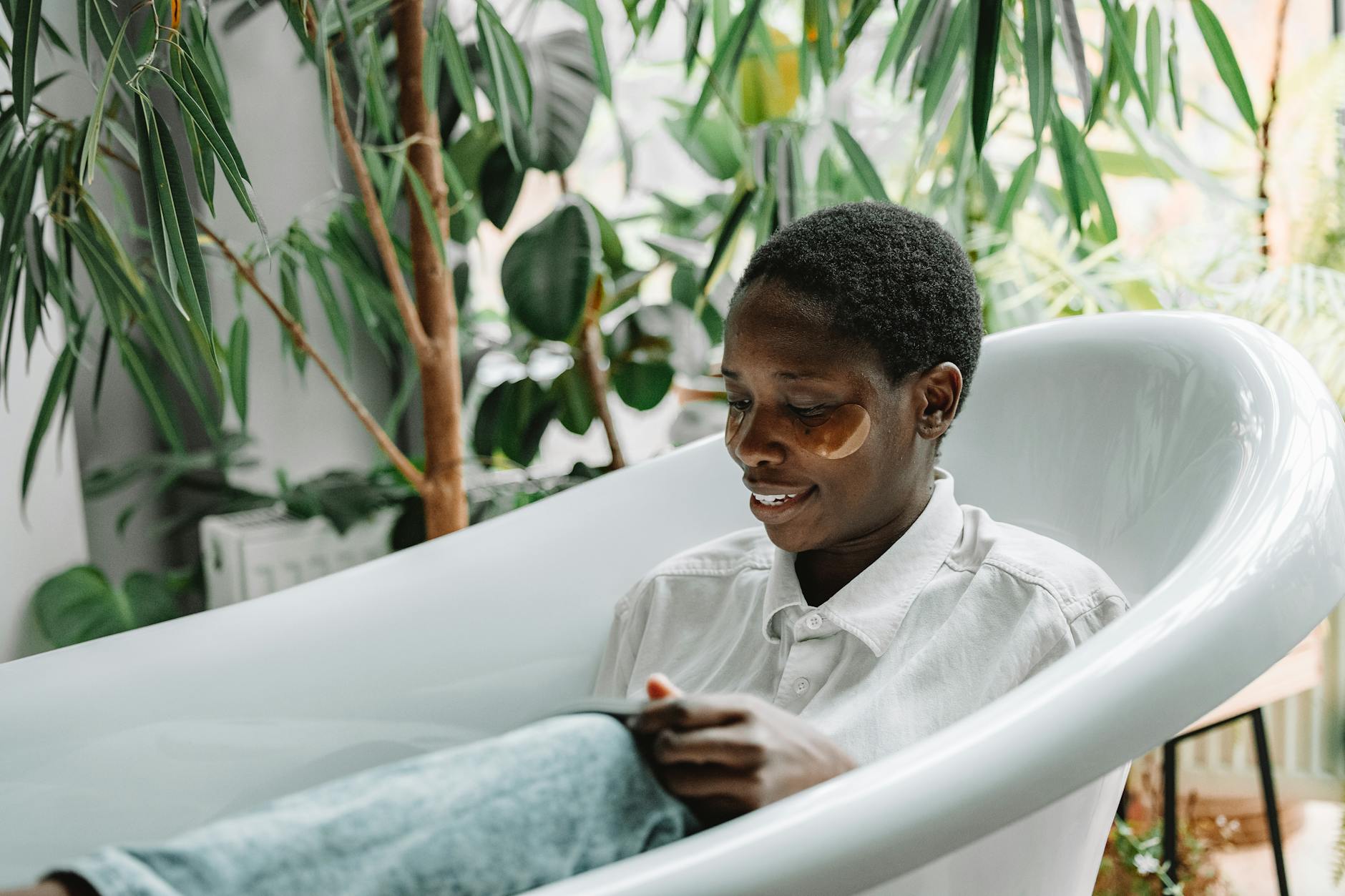 Woman enjoying self-care in a bathtub with under eye patches amidst lush indoor plants. - self care habits