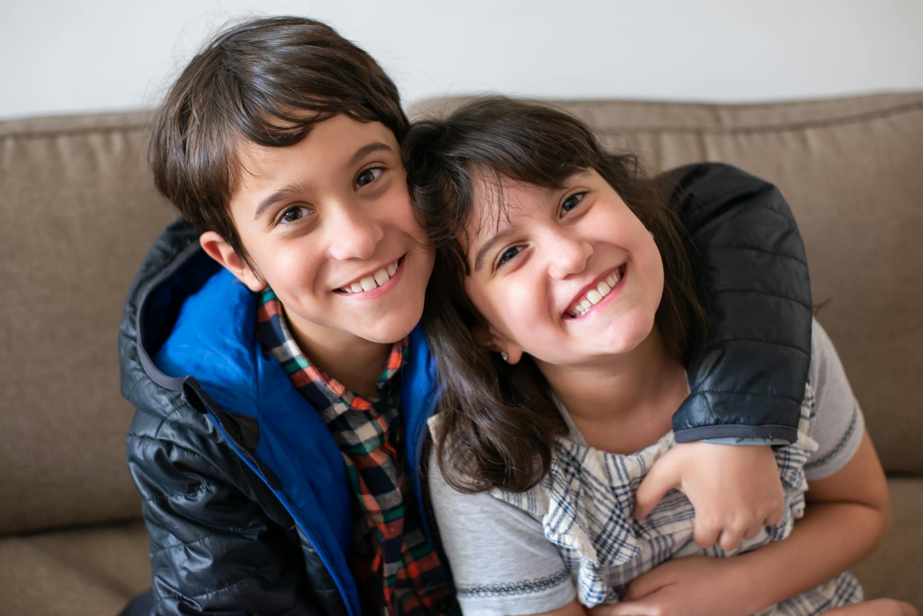 Happy siblings smiling and embracing while sitting on a comfortable sofa indoors. - sibling rivalry causes