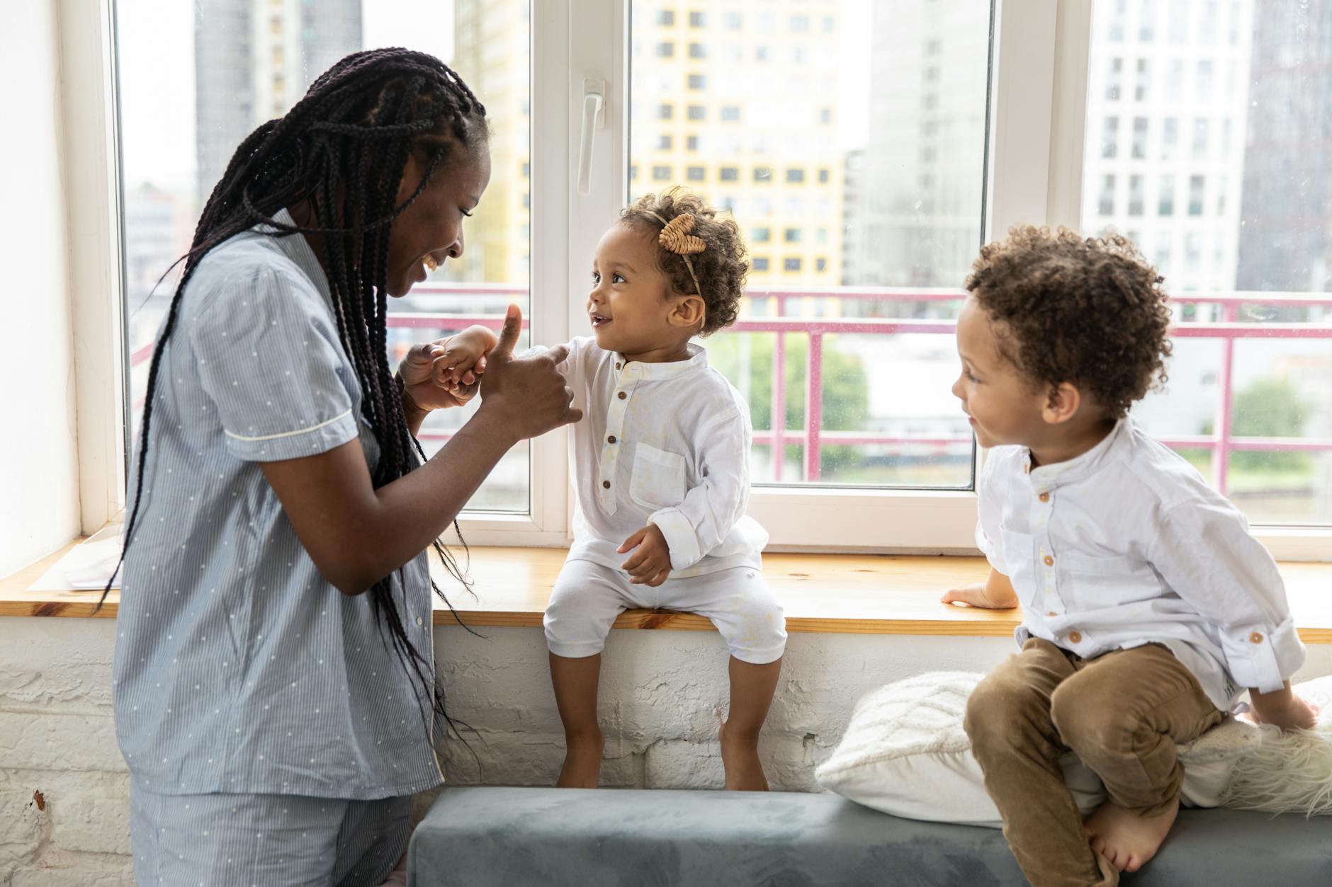 A mother and her young children enjoying playful interaction on a windowsill indoors. - sibling rivalry causes