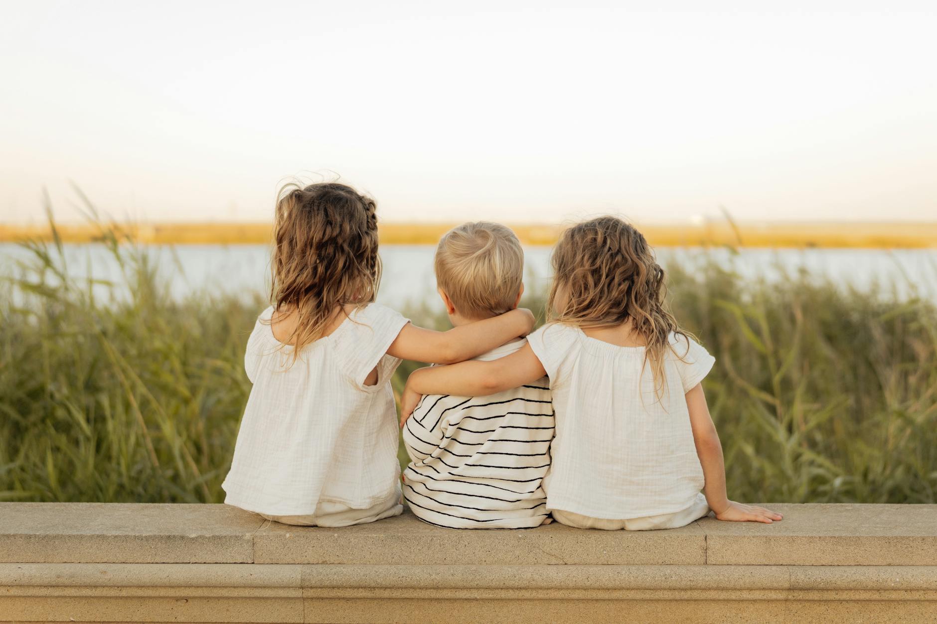 Three children sit on a stone ledge, gazing at a serene lakeside scene, embodying friendship and tranquility. - sibling rivalry definition