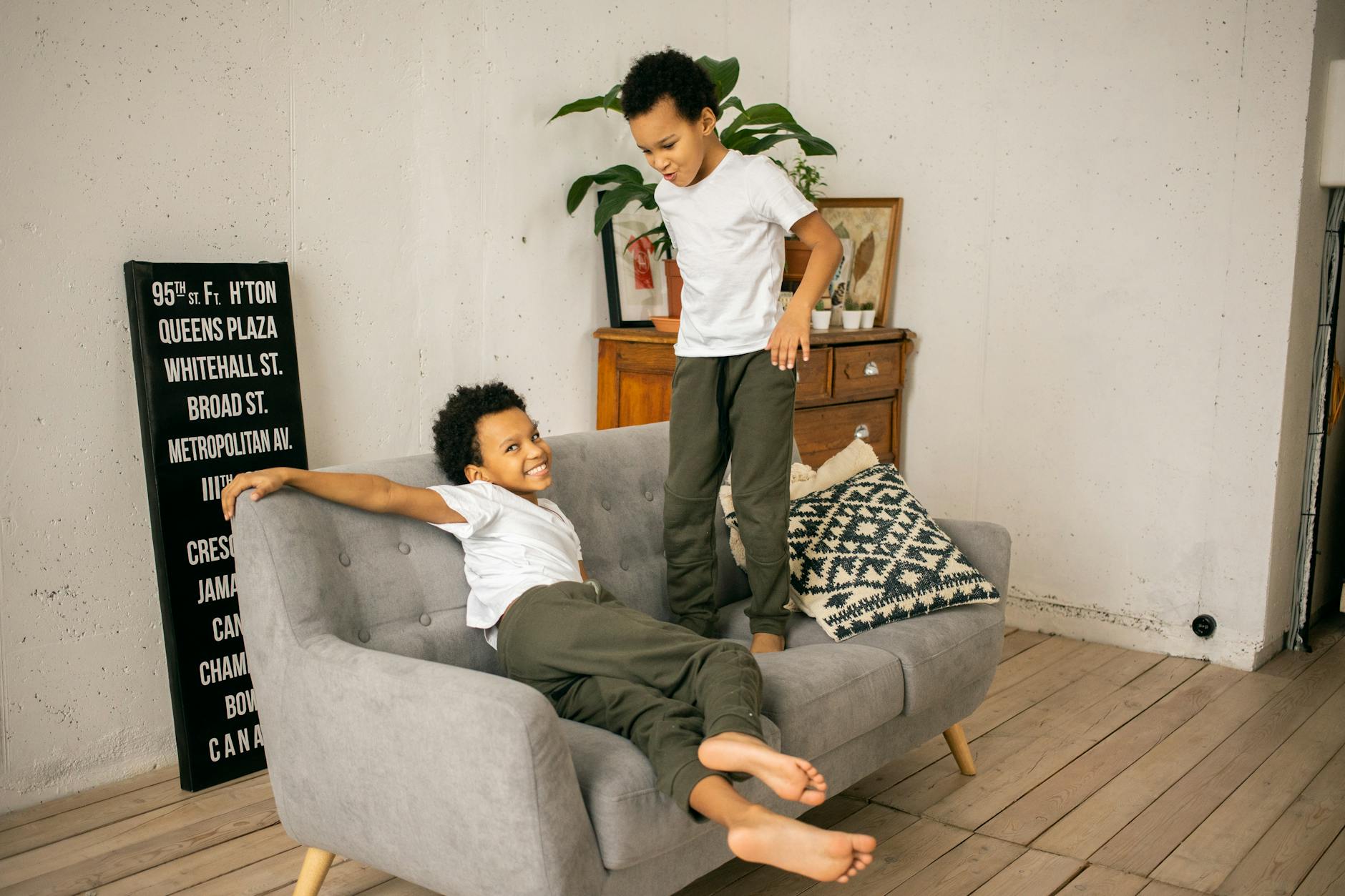 Full body of funny barefoot African American boys playing on comfortable couch in light room with signboard and wooden cupboard - sibling rivalry definition