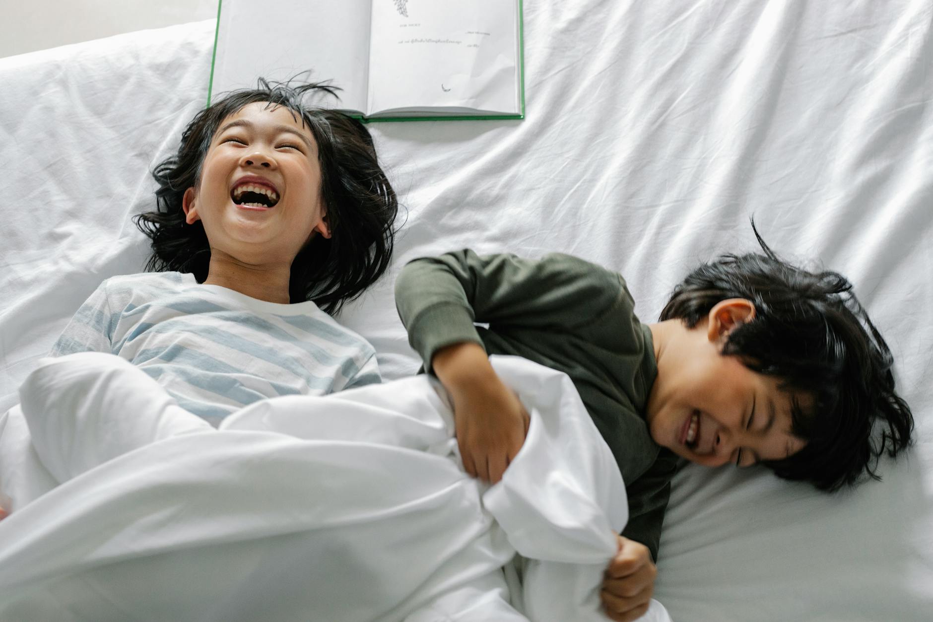 From above of laughing Asian boy and girl lying on bed with book under white blanket in morning in bedroom - sibling rivalry definition