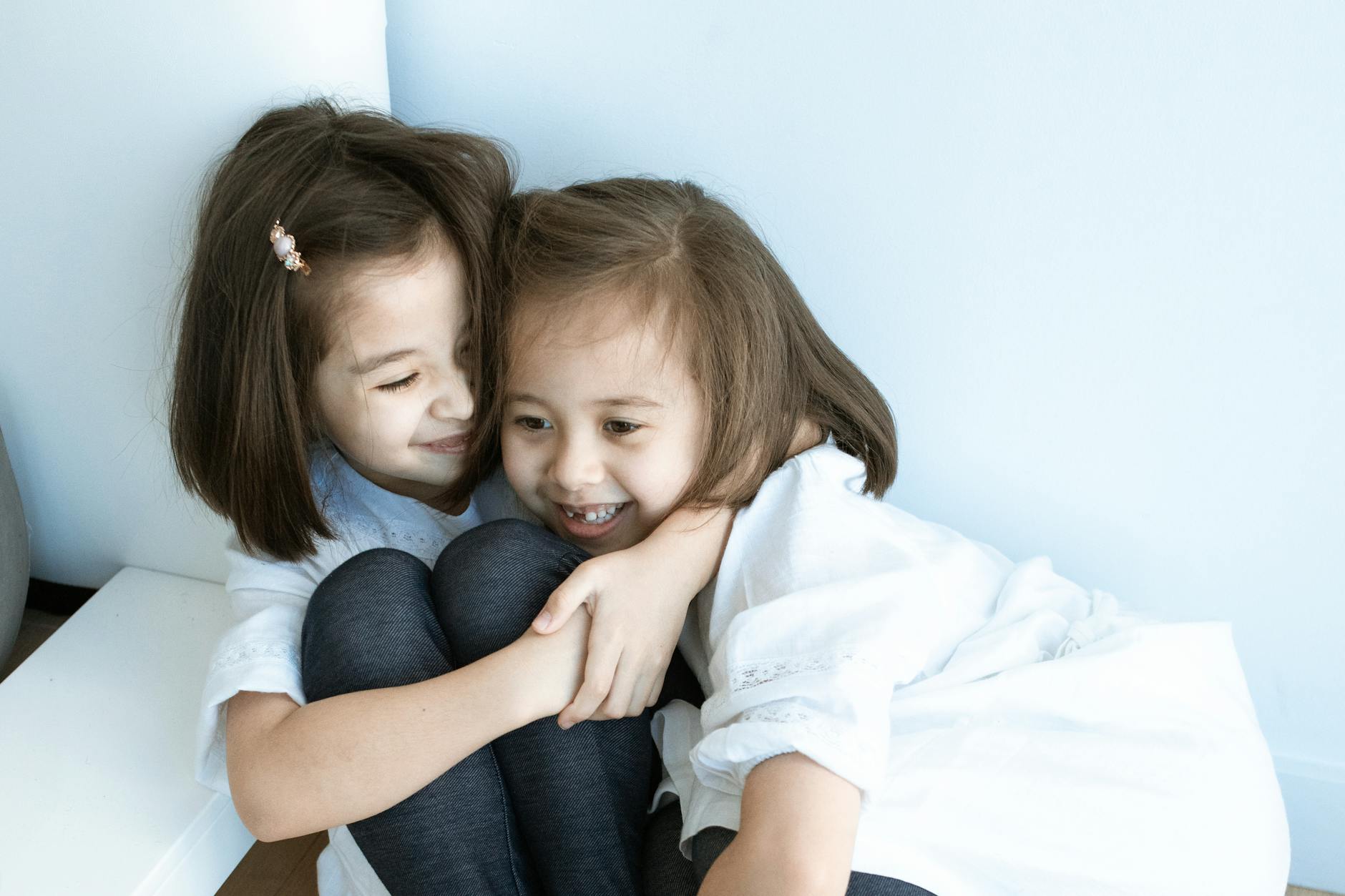Two young girls in white tops embrace joyfully indoors, capturing sibling love and happiness. - sibling rivalry tips