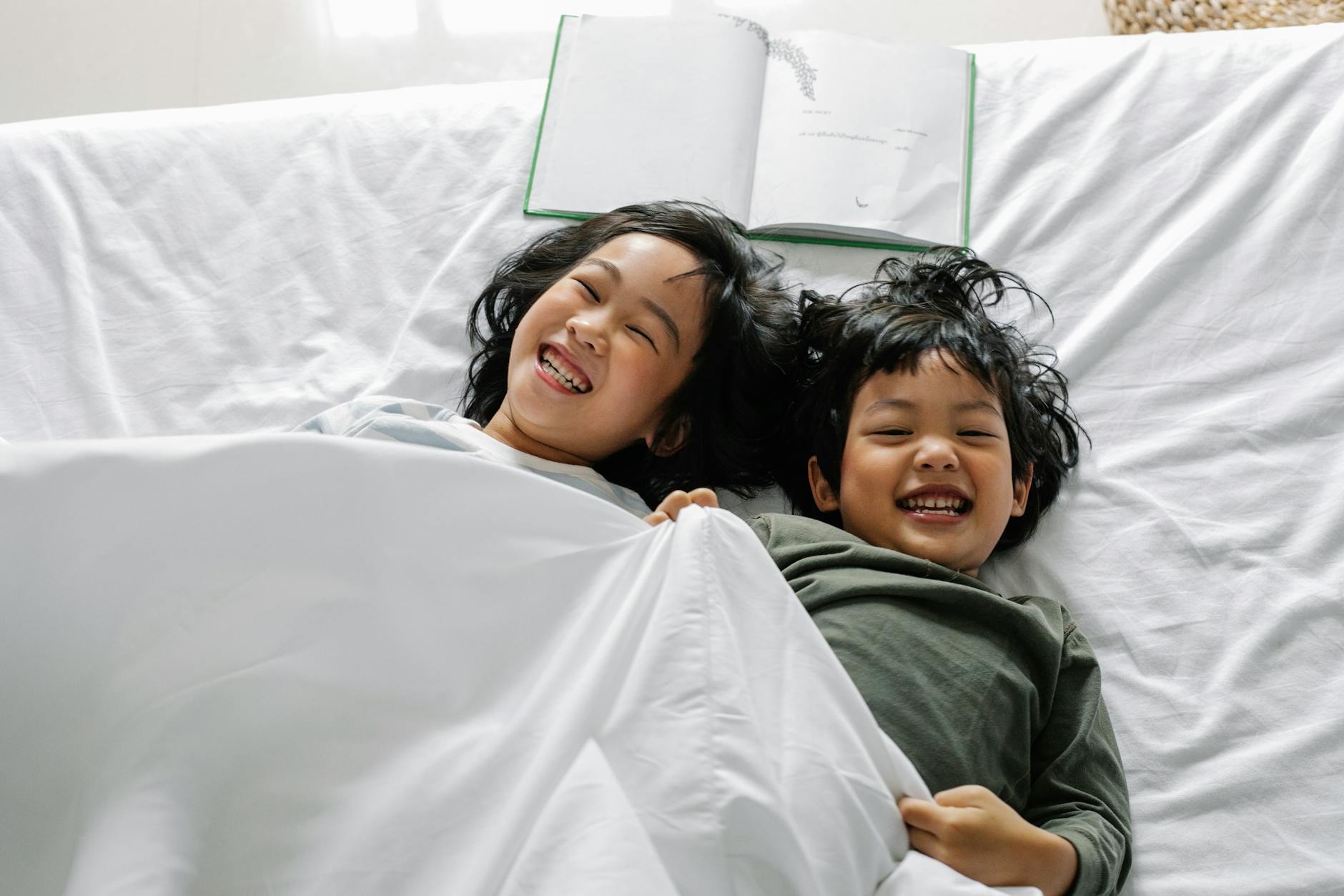 From above of Asian siblings laughing happily while lying in bed under soft blanket - sibling rivalry tips