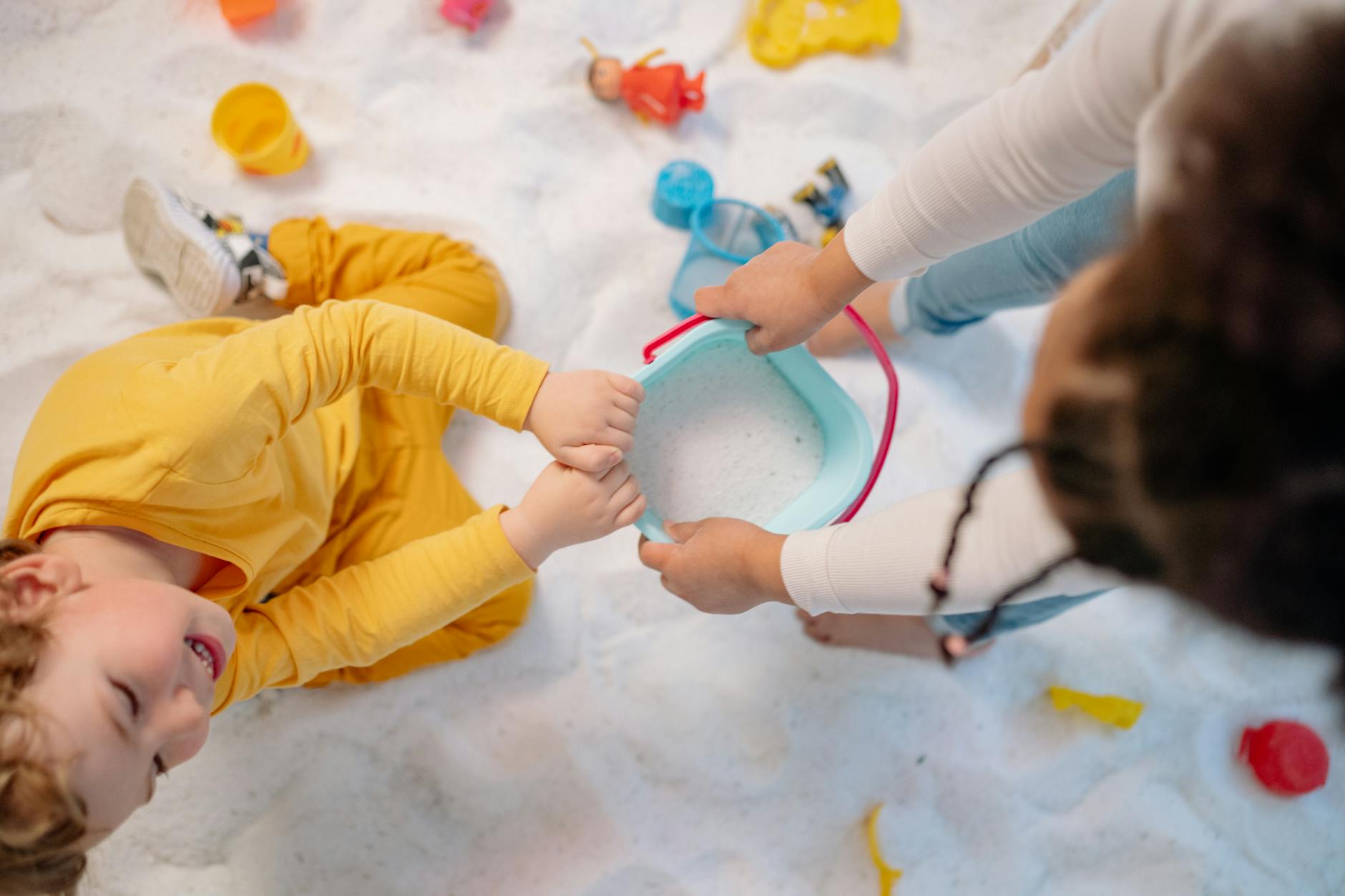 Two children play with toys and a bucket on a sandy surface indoors. - social skills playdates