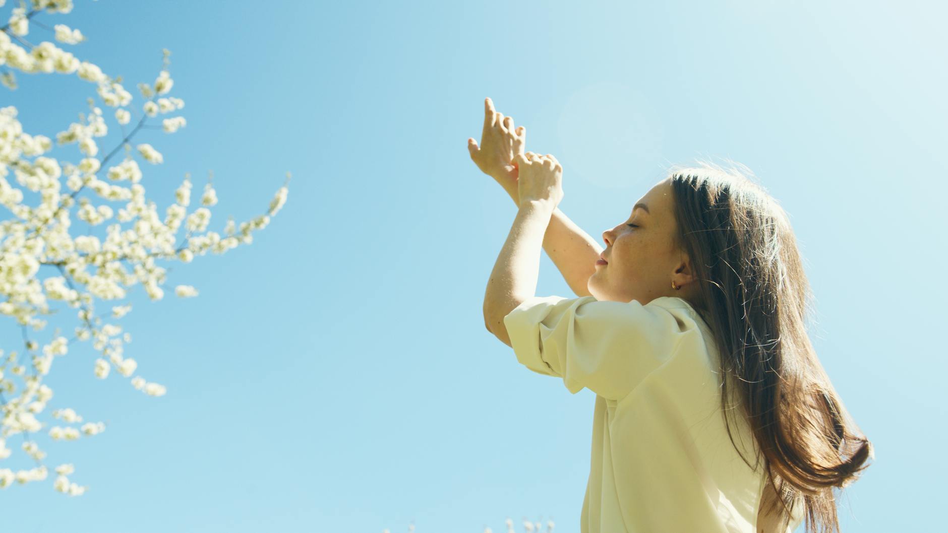 A serene moment with a young woman raising her hands under the clear blue sky filled with blossoming branches. - spring allergy anxiety