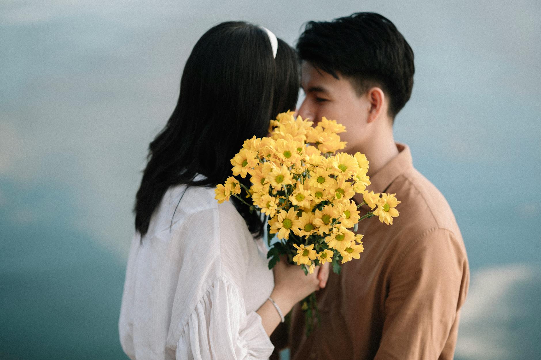 A romantic couple sharing an intimate moment by the water, holding a bouquet of yellow flowers. - spring dating ideas