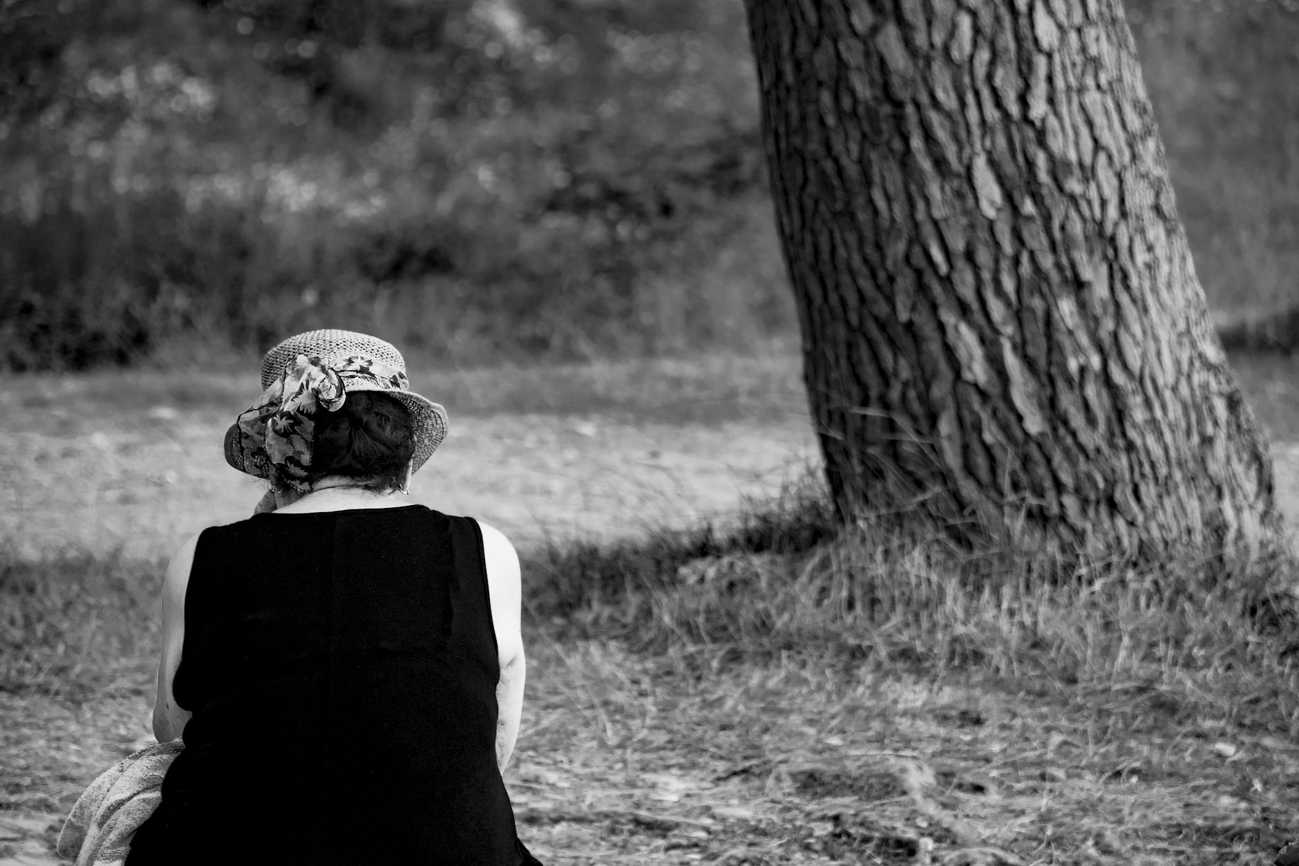 Black and white back view of anonymous female in stylish straw hat sitting on lawn during weekend in summer - spring loneliness