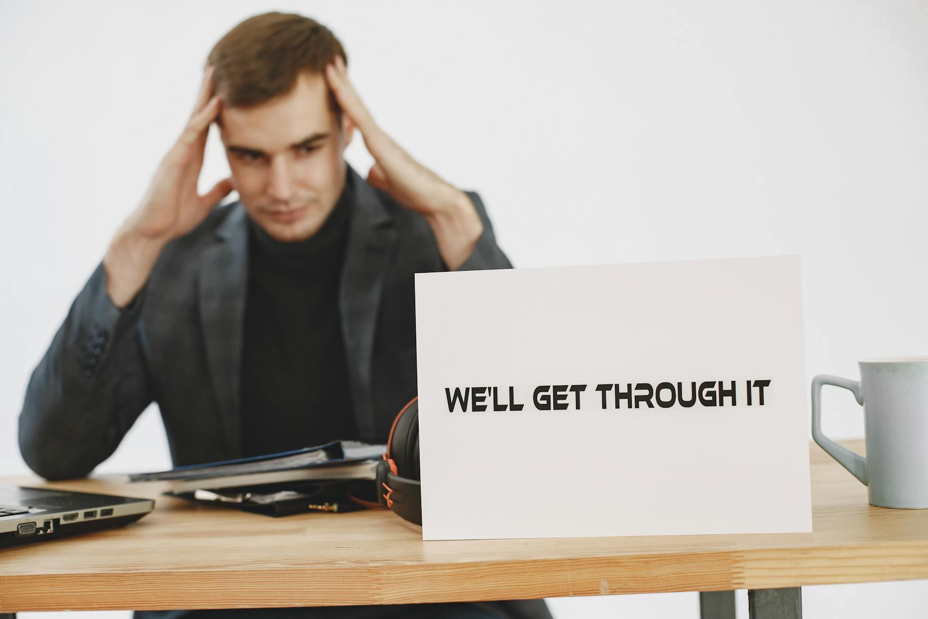 A stressed man sits at a desk with a motivational sign saying 'We'll Get Through It'. - stop overthinking