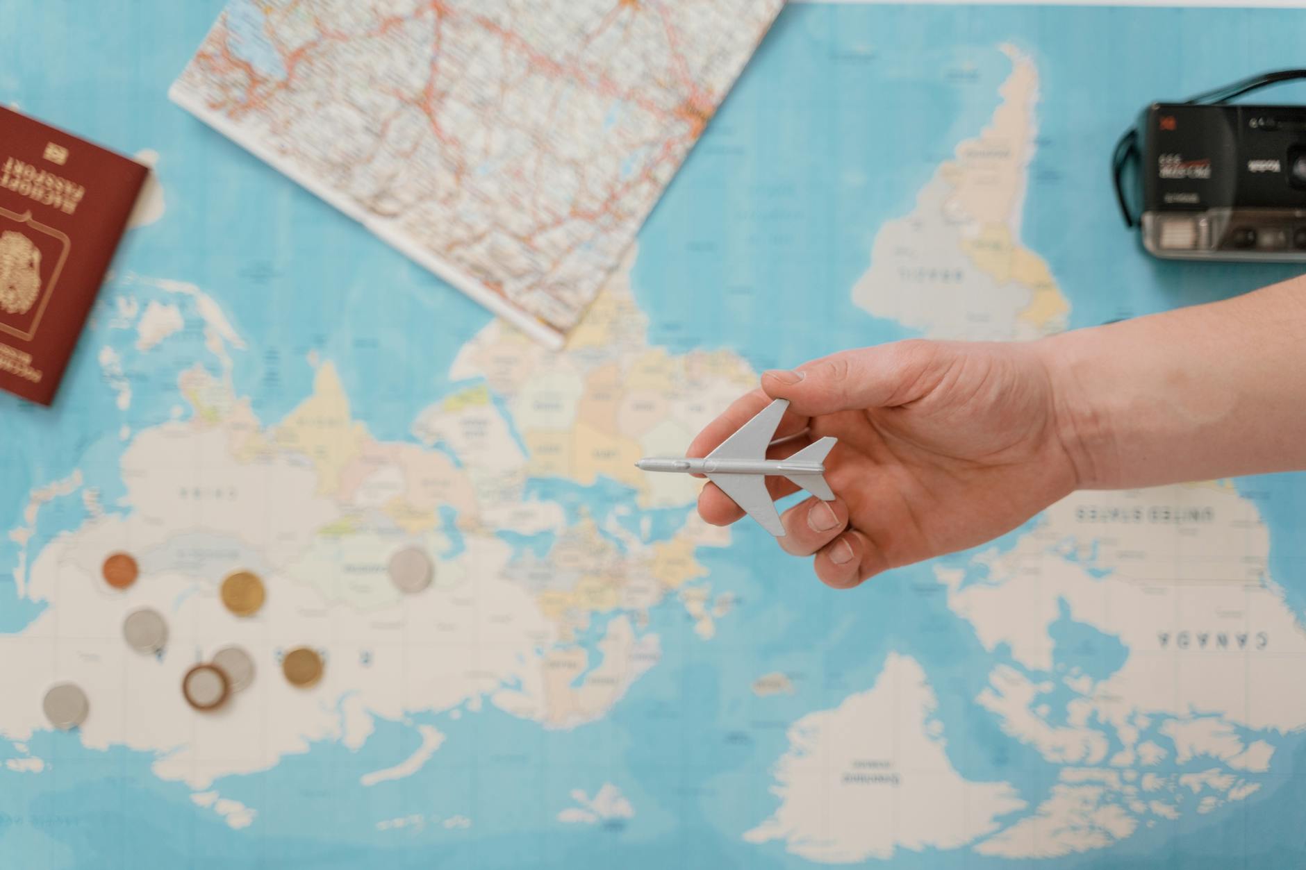Hand holding a toy airplane over a world map, surrounded by coins, camera, and passport. - summer vacation planning