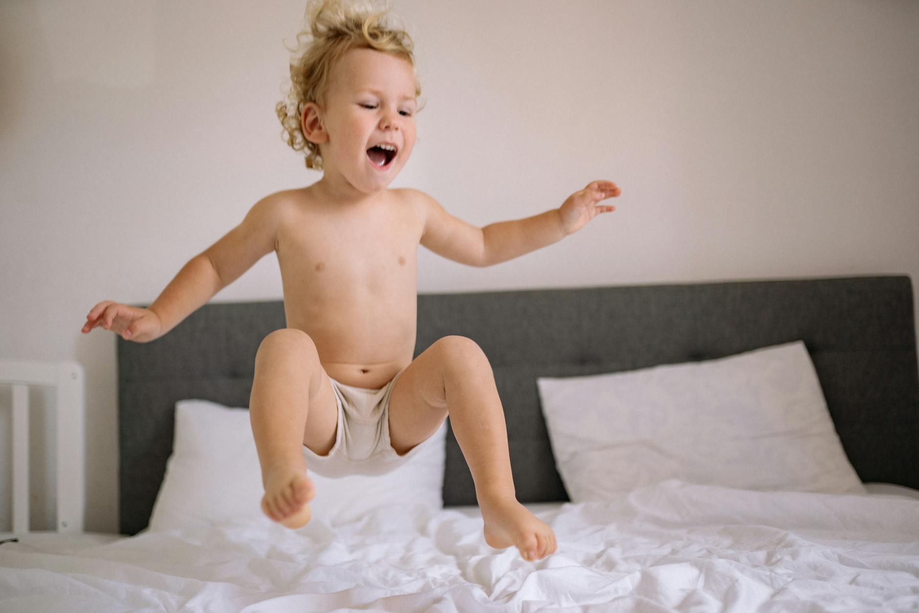 A joyful child with curly hair jumps on a bed indoors, expressing carefree fun and happiness. - toddler energy bursts
