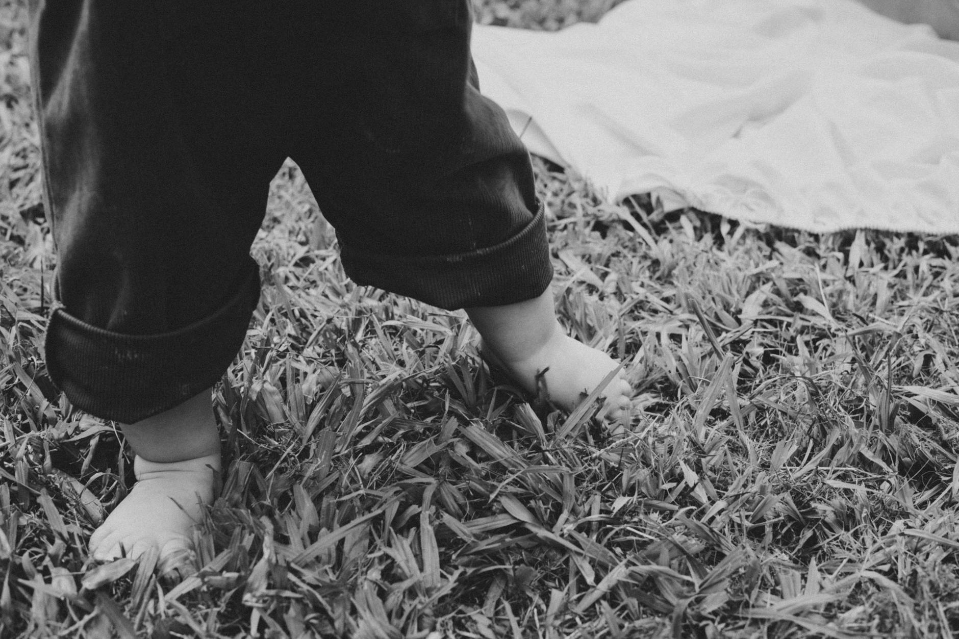 Black and white photo of a baby taking first steps on grass, showing feet and rolled-up pants. - toddler spring nature