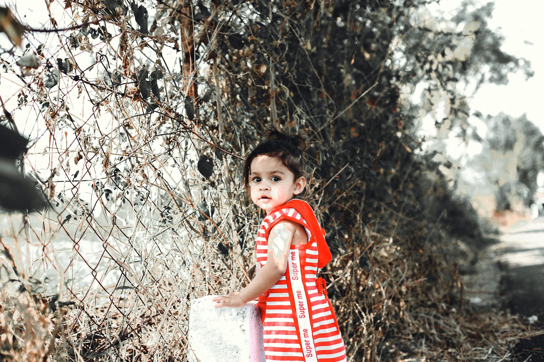A cute child in a red striped dress stands by a fence surrounded by foliage. - toddler tantrums outdoors