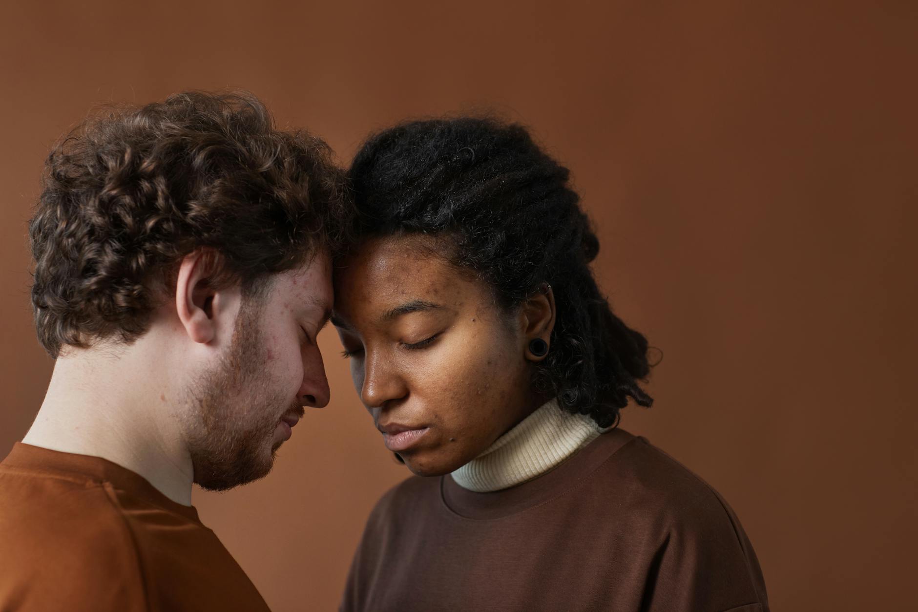 Close-up of an interracial couple embracing with eyes closed against a brown background. - vulnerability in relationships
