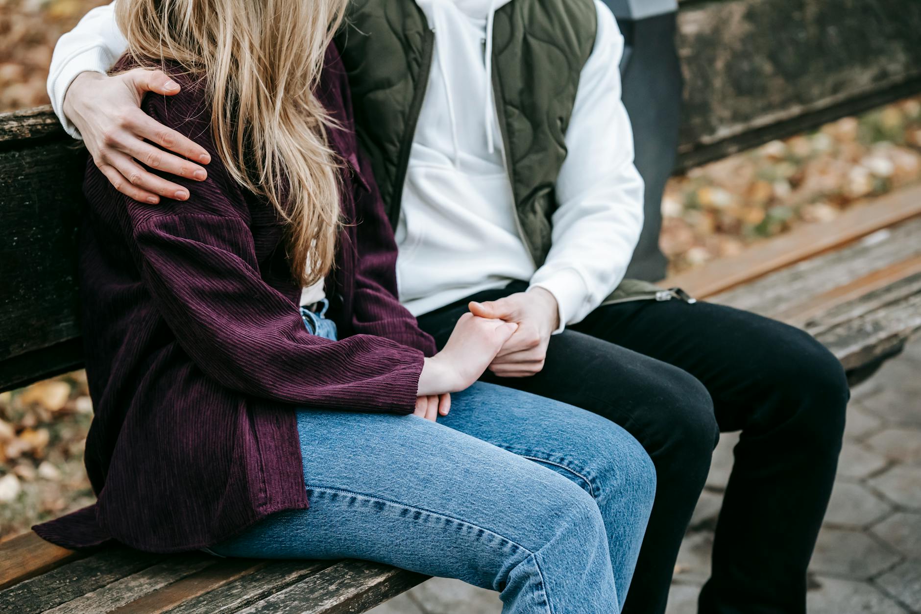 Crop anonymous tender young couple in casual stylish outfit cuddling and holding hands on wooden bench - vulnerability in relationships