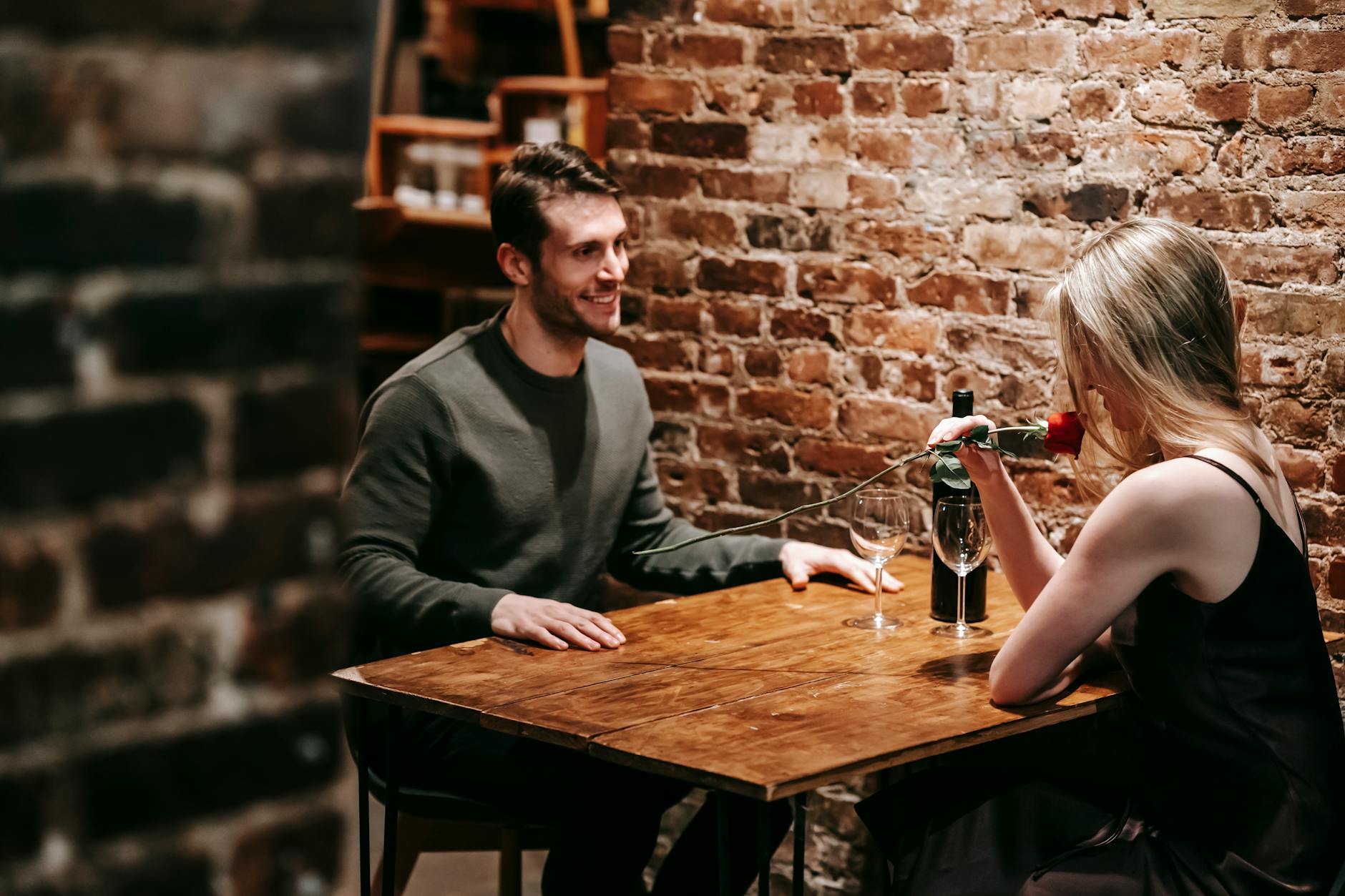 Young couple in elegant clothes sitting in light cafeteria at wooden table near brick wall and having romantic dinner with red rose and wine near glasses - vulnerable dating